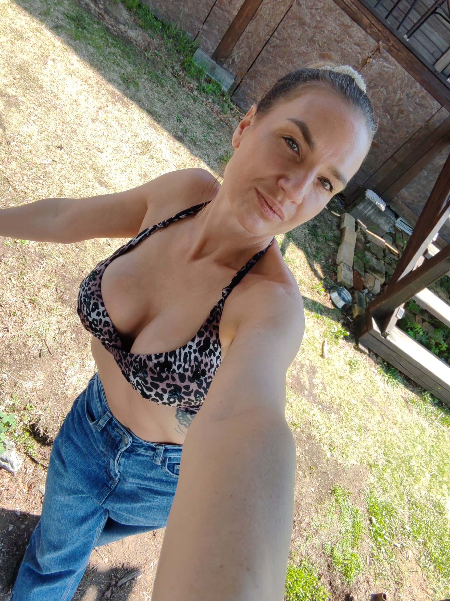 A fair-skinned adult female is depicted in a high-angle selfie taken outdoors in Yelizovo, Russia. She looks directly into the camera with a slight smirk, exhibiting light-colored eyes and natural-toned lips. Her light brown hair is pulled back into an updo. She wears a leopard-print bikini top adorned with dark sequins, revealing significant cleavage. Below the top, a portion of blue denim jeans is visible, partially unzipped/unbuttoned at the waist. A dark tattoo is partially visible on her left side, below the bikini top. The background features a patchy ground cover of dry grass, green grass, and bare dirt, under strong sunlight casting a diagonal shadow from the upper left. To the right, a rustic wooden structure, possibly a railing or support beam, is visible with stones at its base. Above her head, a wooden ceiling or roof made of panels and beams is apparent. A metallic bowl rests on the ground in the mid-right portion of the frame.