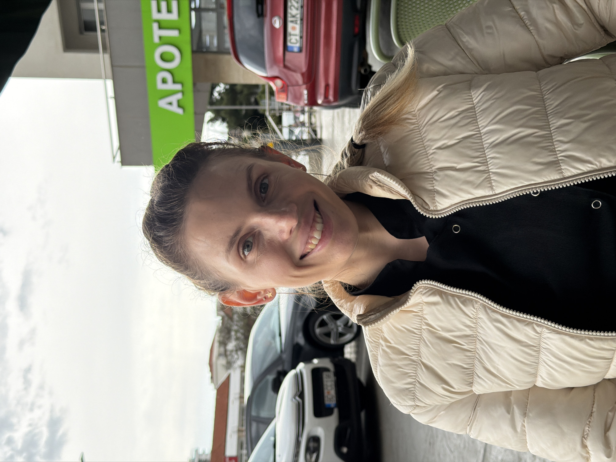 A fair-skinned woman with brown hair pulled back is smiling at the camera. She is wearing a light beige puffer jacket and a black top. Behind her, there is a pharmacy storefront with the word "APOTEKA" in large green letters. A red car with a license plate reading "CT AK22" is parked behind the pharmacy. Other vehicles are visible in the background. The setting appears to be a street in Budva, Montenegro.