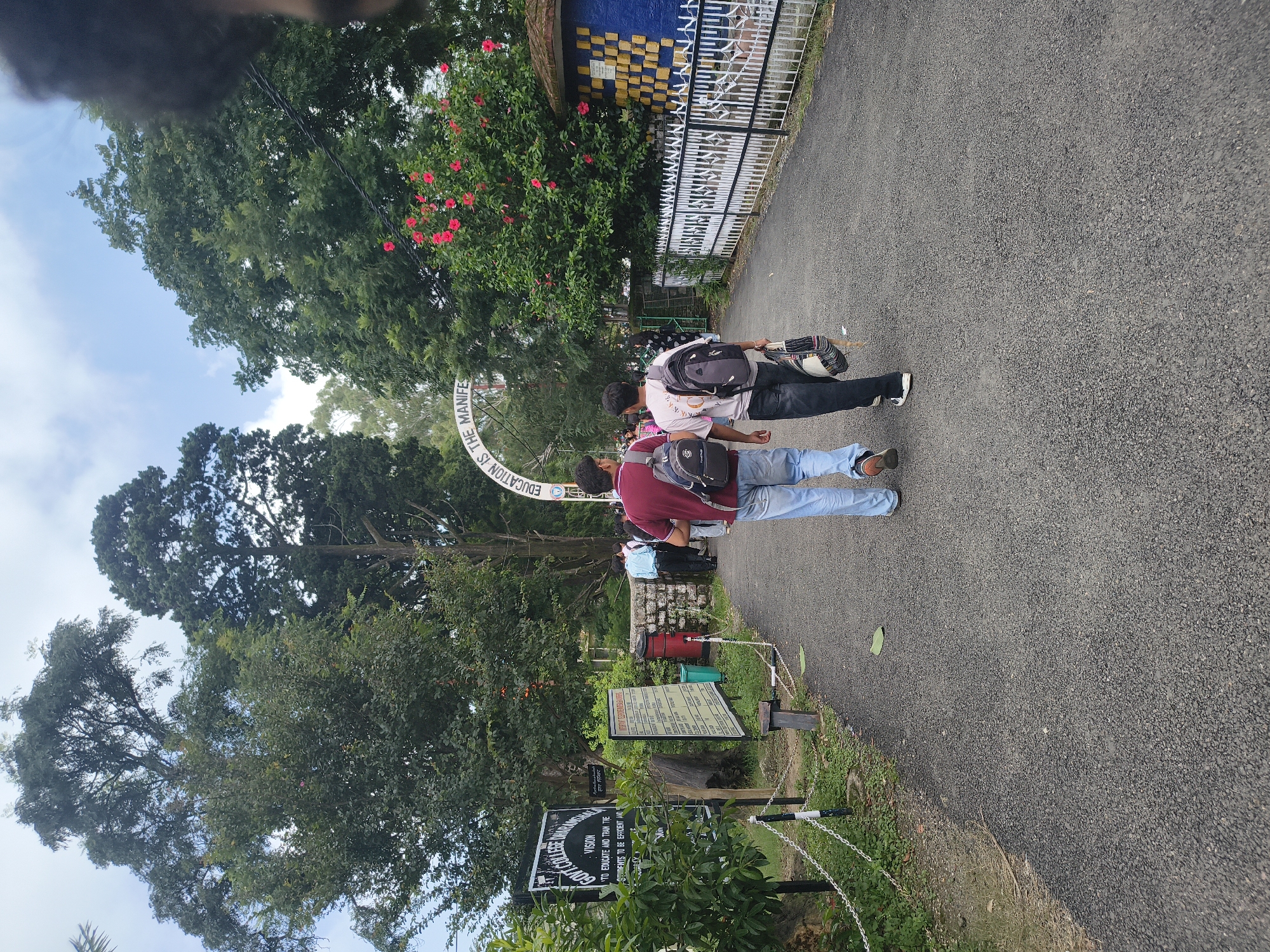 The image depicts an outdoor scene on a paved pathway, likely on the grounds of Govt. College Dharamshala, India. The pathway is flanked by lush green trees and foliage. On the left side of the path, a black sign with white text, which reads "GOVT. COLLEGE DHARAMSHALA", is visible. Further down the path, an arched gateway with text "EDUCATION IS THE MANIFEST" is present. Two young men, each carrying a backpack, are walking away from the camera along the pathway. One is wearing a maroon t-shirt and blue jeans, while the other is in a light-colored t-shirt and dark pants, carrying a woven bag. A white fence with decorative patterns runs along the right side of the path, behind which is a wall with a mosaic of blue and yellow squares. The sky is overcast with scattered clouds. The overall atmosphere is that of an educational institution's campus on a typical day.