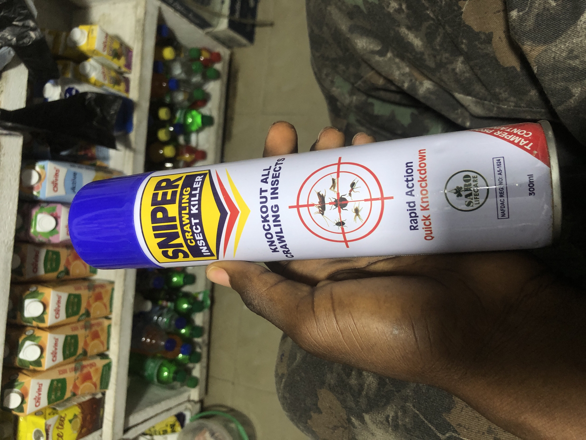 A person's dark-skinned hand holds a can of "SNIPER CRAWLING INSECT KILLER" in a store. The can is predominantly white with blue, yellow, and red text and graphics. The label features a crosshair target with images of insects, text indicating "KNOCKOUT ALL CRAWLING INSECTS," and "Rapid Action Quick Knockdown." Below this, a circular logo reads "SARO LIFECARE" with a registration number. The background shows shelves stocked with various beverages, including juice cartons and bottled drinks. The store appears to be in Ado, Nigeria, based on the presence of NAFDAC registration. The person is wearing camouflage-patterned clothing. The lighting is artificial, suggesting an indoor setting.