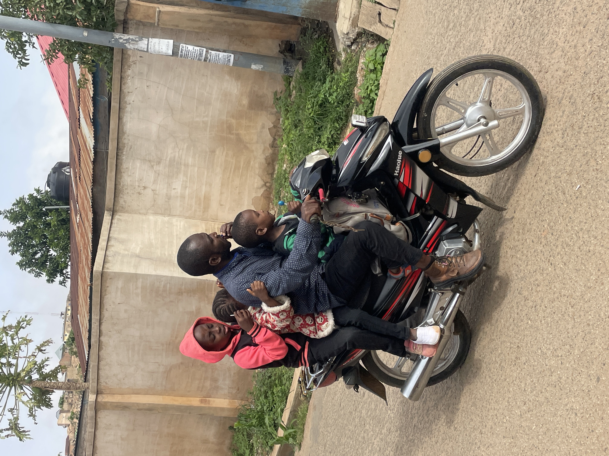 A man is riding a black Haojue motorcycle on a paved road in Jos, Nigeria. Two children are seated in front of him, one between his arms, while another child is seated behind him. The child in front wears a green shirt, and the child behind wears a pink hooded jacket. The man wears a blue patterned shirt and black pants. The child behind the driver appears to be eating. Buildings with corrugated metal roofs are visible in the background, as is a light pole with affixed notices. Green foliage grows near the building and along the roadside.