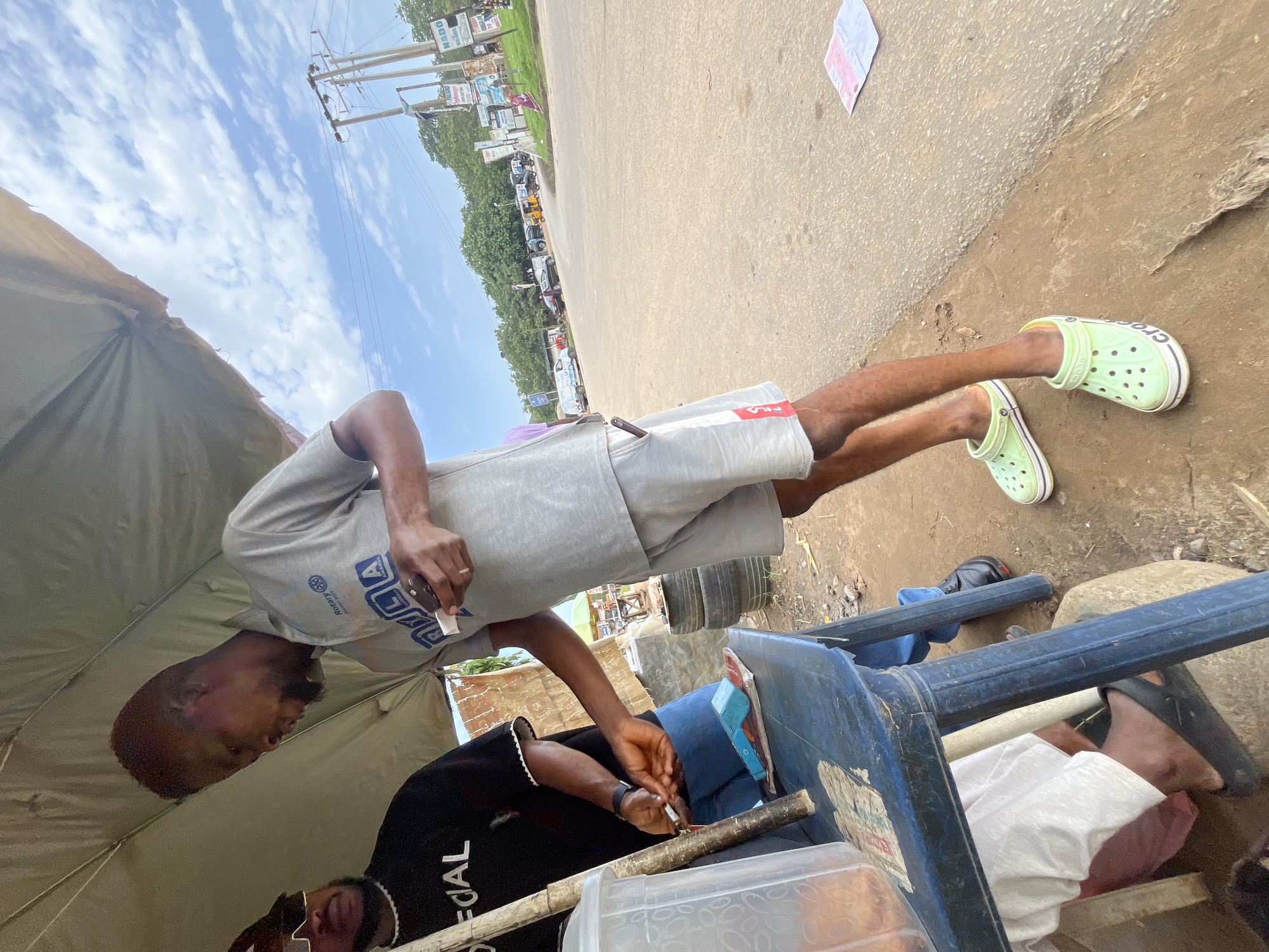 In Abuja, Nigeria, under a makeshift canopy, two individuals are engaged in an interaction near a roadside stall. The person on the right, wearing a grey polo shirt with "Rotary" and "ACLAWARDS" logos and grey shorts, is holding a small white object and appears to be smoking. Their feet are clad in light green Crocs. To the left, another individual in a black t-shirt with "OFFICIAL" printed on it, and sunglasses, is attending to a plastic container and a wooden pole, possibly serving a customer. A blue plastic table is present, with various items on it, including a turquoise handheld device and papers. The background shows a dusty road, scattered tires, and distant trees and utility poles, suggesting an outdoor commercial setting in a Nigerian city.