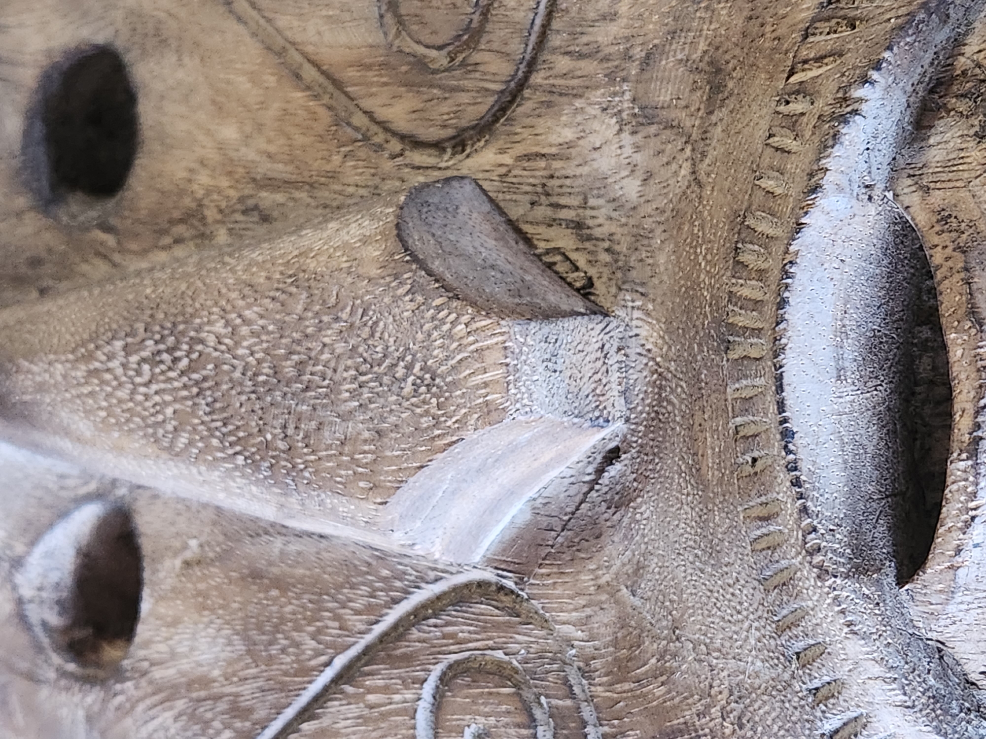 The image is a close-up, high-angle shot of a carved wooden mask. The mask is detailed, with visible wood grain and textured surfaces. The prominent features are the eyes, nose, and mouth. The eyes are dark, hollowed-out openings. The nose is broad and appears to have flared nostrils carved into it. The mouth is an open cavity with intricate carved details around the rim, resembling teeth or a decorative border. Swirling decorative lines are present on the cheeks, framing the central facial features. The overall color palette is a range of earthy browns and grays, suggesting aged wood. There is no discernible background or environmental context visible in the frame. The image does not provide any information about a specific location in Maitland, United States of America, nor does it depict any people, actions, or interactions.