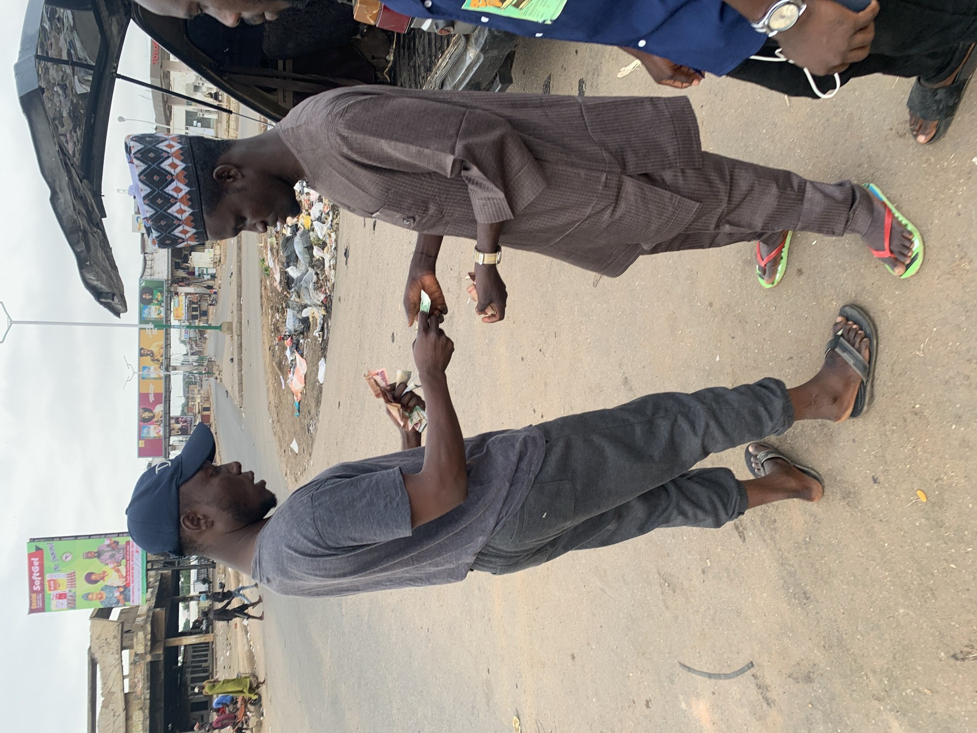 Two men are exchanging money on a street in Jos, Nigeria. One man, wearing a dark brown traditional garment and a patterned kufi cap, passes money to a second man wearing a blue baseball cap and a grey t-shirt. The exchange takes place near an open car trunk. The surroundings appear to be an urban street with visible litter, billboards, and nearby buildings.