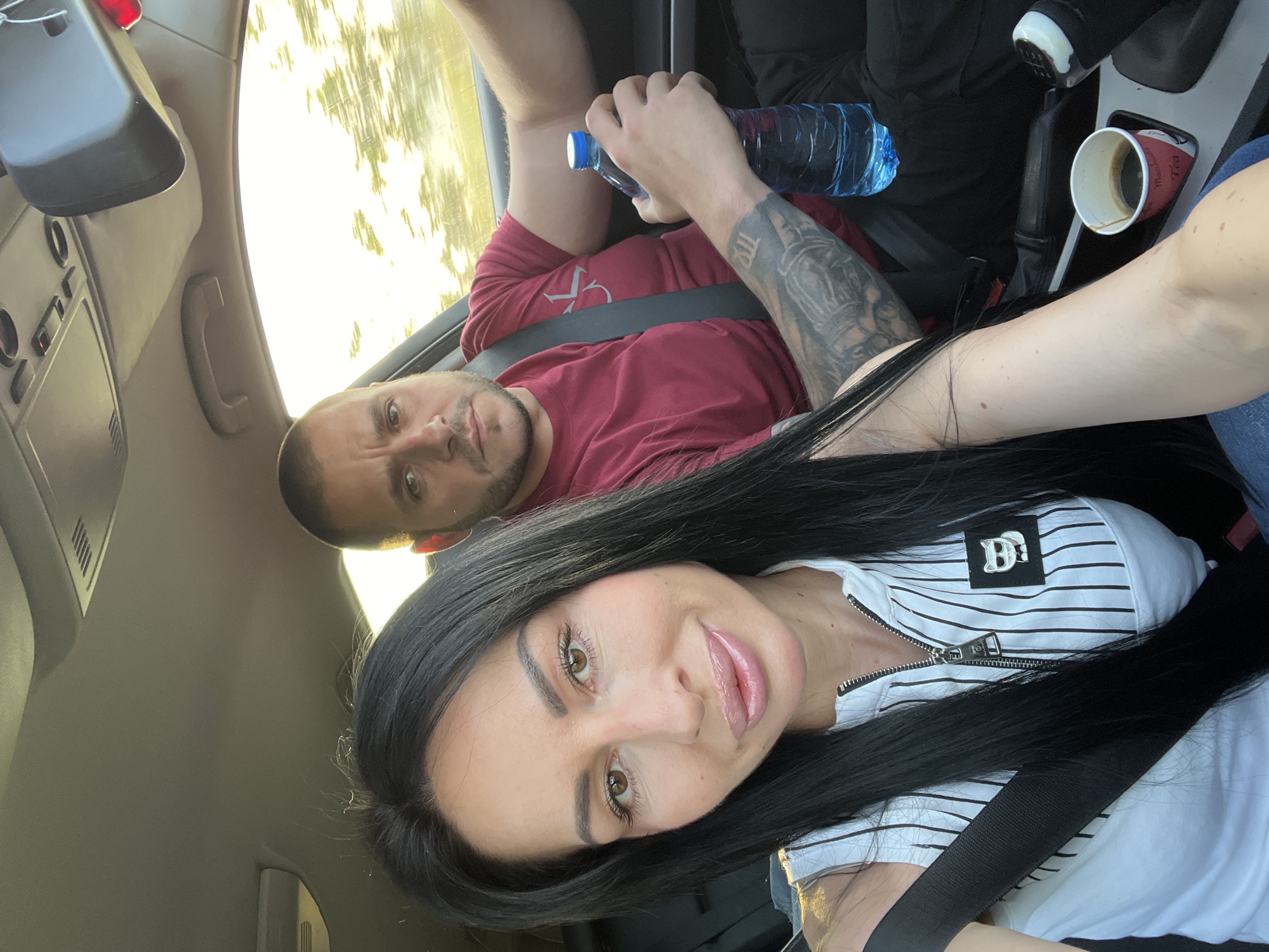 This image is a selfie taken inside a car, featuring two individuals, likely a couple, in Bogomilovo, Bulgaria. The shot is tilted, with the subjects appearing diagonally.
In the foreground, a woman with long, straight black hair, light brown eyes, and light pink lip gloss looks directly at the camera with a slight smile. She is wearing a white top with thin black horizontal stripes, a prominent silver zipper down the front, and a silver square emblem on her chest featuring the letter "D" (and possibly another intertwined letter or design). She is secured by a black seatbelt.
Behind and to her left, a man with a short, dark beard and shaved head, also with light brown eyes, looks towards the camera. He wears a solid red long-sleeved shirt and holds a blue plastic water bottle in his left hand. His left arm is heavily tattooed with a dark sleeve design. He is also wearing a black seatbelt.
The interior of the car is visible, including parts of the dashboard, a sun visor grab handle, and a rear-view mirror. To the right of the man, on the center console, a disposable coffee cup, partially filled with coffee, is visible. The cup has "MADE FOR YOU" and "TEA" printed on its side. Bright natural light streams through the car windows, suggesting it is a sunny daytime, possibly late morning or afternoon. The bright light also reflects off the car's exterior, visible through the windows. The overall impression is one of travel or a road trip.