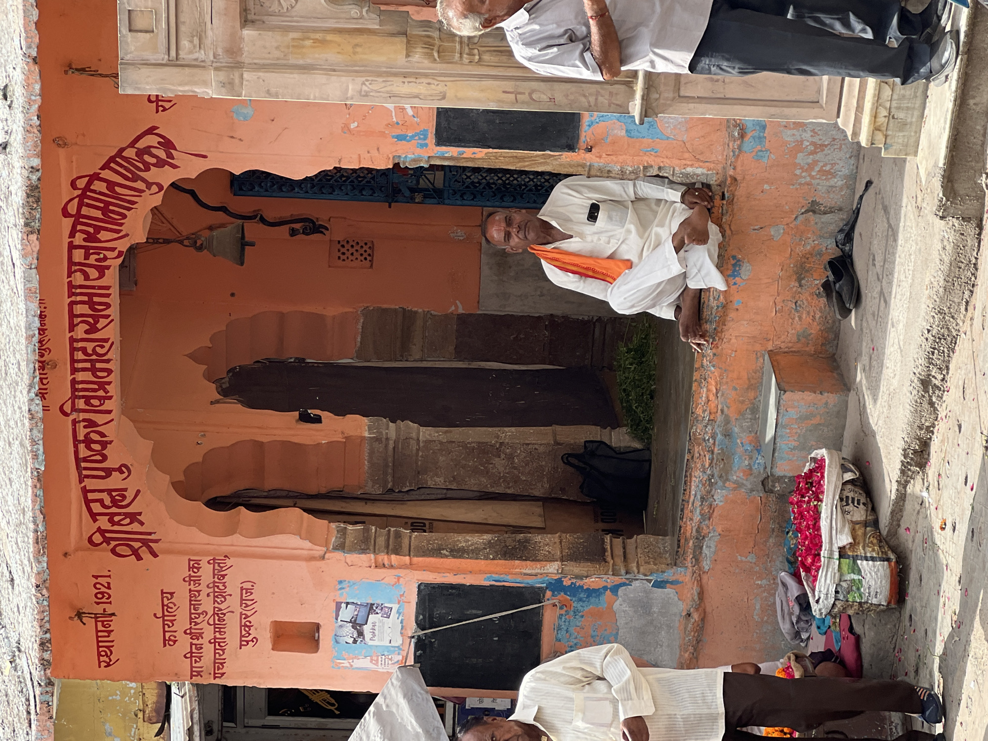 A man wearing a white kurta and pants sits on a ledge outside an orange building in Pushkar, India. He has an orange cloth draped over his shoulders. In front of him is a black bag and a small pile of green leaves. To his right, another man in a light gray shirt and dark pants stands on steps.
The building has Hindi text in red paint above an arched doorway. The text mentions "Sthapana - 1921" and "Shree Brahma Pushkar Mahasamaj Samiti". Below this, more Hindi text reads "Karyalaya Prachin Shri Raghunath Ji ka Panchayati Mandir Choti Basti, Pushkar (Raj.)". A small bell hangs by a chain to the left of the doorway.
To the left of the men, a man in a white striped shirt and brown pants stands, partially visible. In front of him, a large woven bag filled with red flowers is on the ground. The ground is paved with concrete and scattered with rose petals. The environment appears to be a street or marketplace.