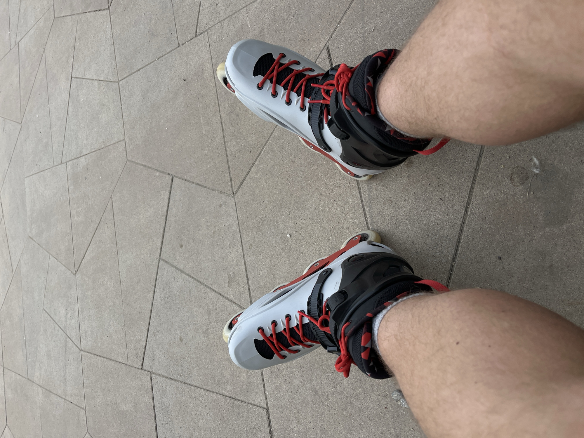 The image shows a first-person perspective of a person wearing rollerblades on a tiled pathway. The rollerblades are white and black with red laces and accents. The person's legs, which are hairy, are visible. The pathway is made of large, irregular grey tiles with dark grout lines. The scene appears to be outdoors, possibly in a park or public space. There is no clear indication of the city of Krasnoyarsk, Russia.