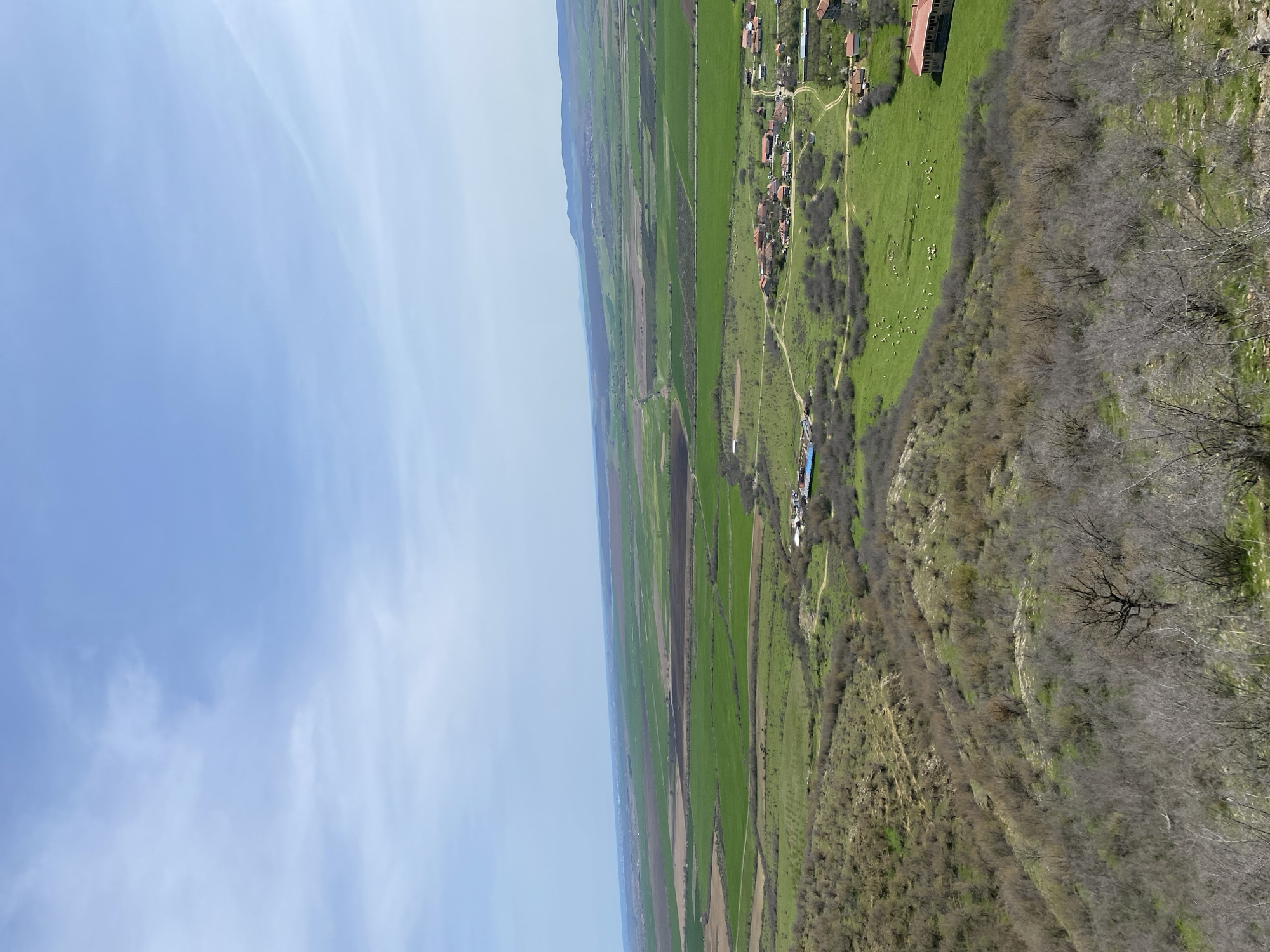 The image presents an aerial view of the landscape in the city of Goritsa, Bulgaria. The sky occupies the upper portion of the frame, exhibiting light blue hues with faint clouds. Below, a landscape unfolds featuring cultivated fields. Patches of green grass are interspersed with brown soil, suggesting agricultural plots or fallow land. Further into the landscape, a rural settlement is observed, characterized by buildings with red rooftops. The village is connected by pathways and roads. Along the right side of the frame, vegetation transitions from green to brown. Some bare trees suggest a wooded area. The transition from field to hillside is marked by clusters of trees and shrubs.