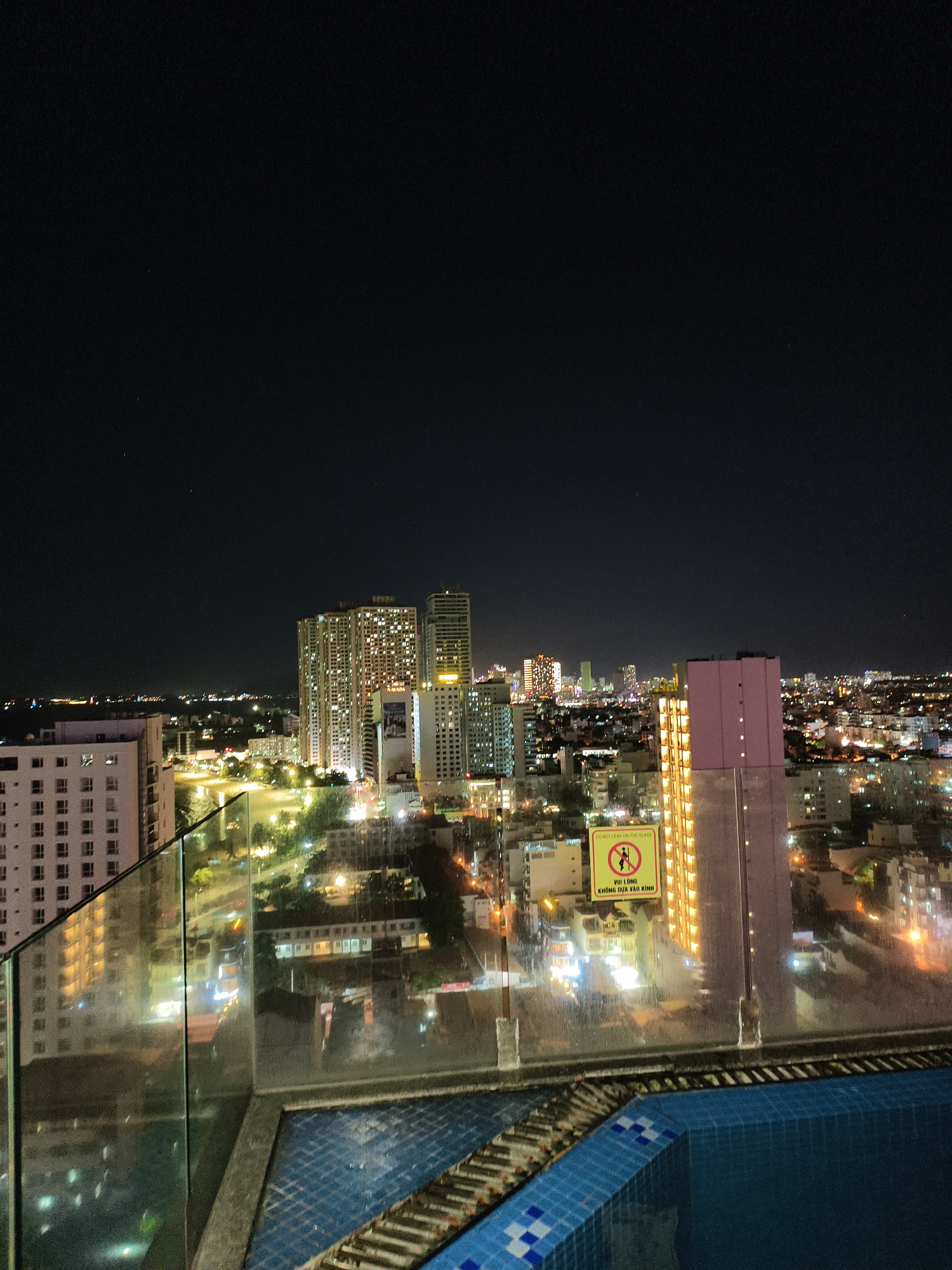 Captured at night in an urban environment, the image showcases a city skyline from an elevated vantage point, likely a rooftop or high-rise balcony. Several brightly lit buildings dominate the middle ground, including apartment buildings with distinct architectural features and possibly a hotel. A pool is visible in the foreground, with a glass barrier reflecting some of the city's lights. A sign with an image of someone leaning on the glass and text below it, in what could be Vietnamese, is prominently displayed. The sky is dark, sparsely dotted with stars. The overall mood suggests an urban, nighttime scene with the potential for leisure or observation.