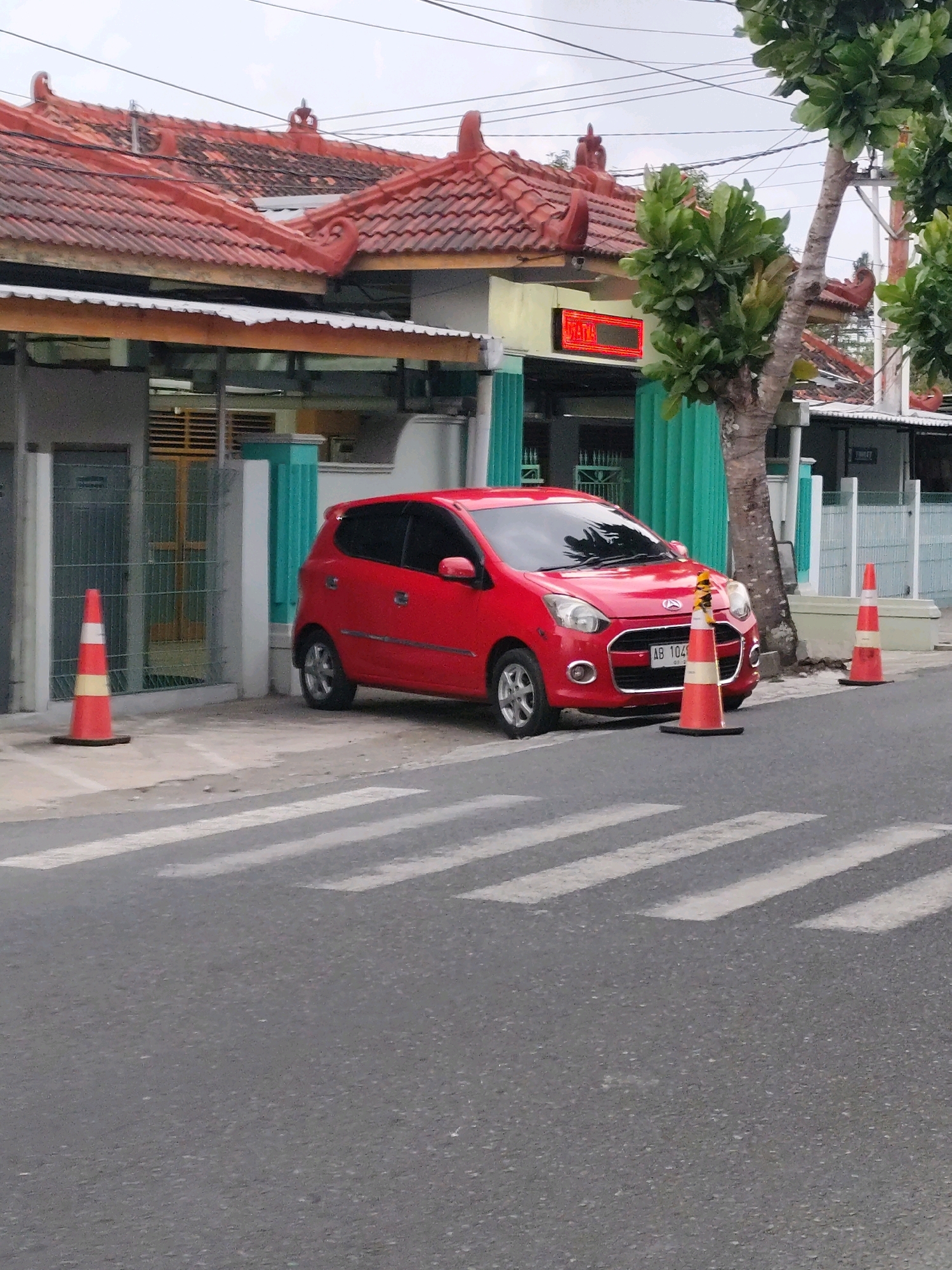 In the city of Bambang Lipuro, Indonesia, a red hatchback car is parked on the side of a road, in front of a building with a red tile roof. A crosswalk is visible in the foreground, with two orange traffic cones placed on the road. The building appears to be a business or office, indicated by a red digital sign displaying "UTAMA". Trees and power lines are also present in the environment, suggesting an urban or suburban setting. The sky is overcast, and the overall lighting suggests it is daytime.