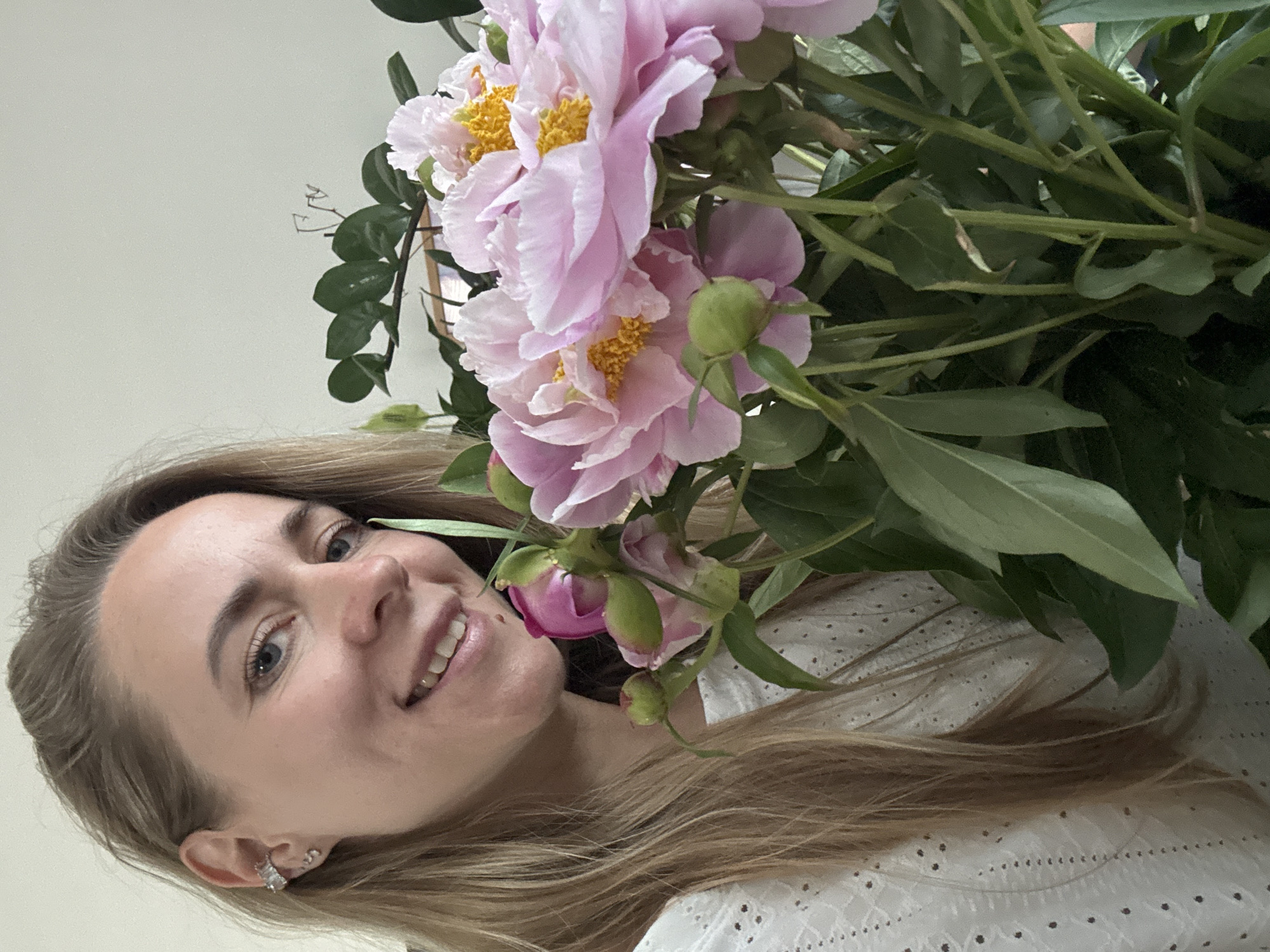 A medium shot portrait of a blonde woman holding a bouquet of pink peonies. She is smiling and looking at the camera. The woman is wearing a white eyelet top and has blonde hair. She has several piercings in her left ear. The background is a plain, light-colored wall. The peonies are large and in full bloom with yellow centers. The bouquet includes several buds and green leaves. The image appears to have been taken indoors. There is no indication of the location being in Chaikovsky, Russia.