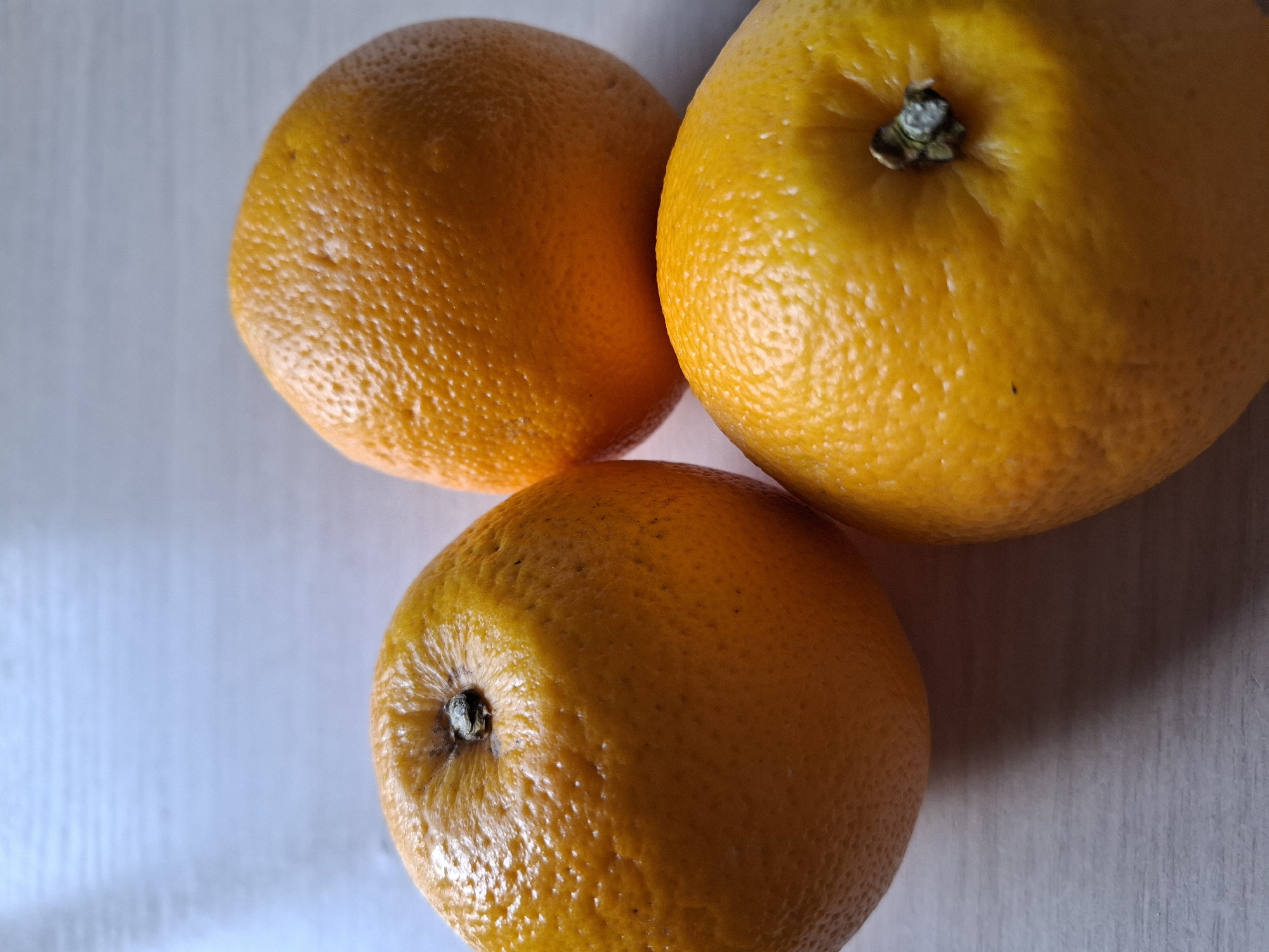 Three oranges are arranged on a light-colored, textured surface, possibly a wooden table. The oranges are positioned in a triangular formation, with one in the foreground at the bottom right, one slightly behind and to the left, and the third to the upper right. The oranges are bright orange with visible peel texture. The lighting creates shadows on the surface, indicating a light source from the upper left. There is no indication of the city of Tyumen, Russia, or any human presence in this image.