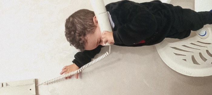 A boy in a black jacket is sprawled across a light gray or beige surface, holding a white telephone receiver to his ear. A coiled white cord stretches from the receiver to a wall-mounted box. The boy’s head is tilted, and his curly hair is visible. To the right of the boy is a circular, decorative white object with cut-out shapes. The scene appears to be indoors against a plain wall.