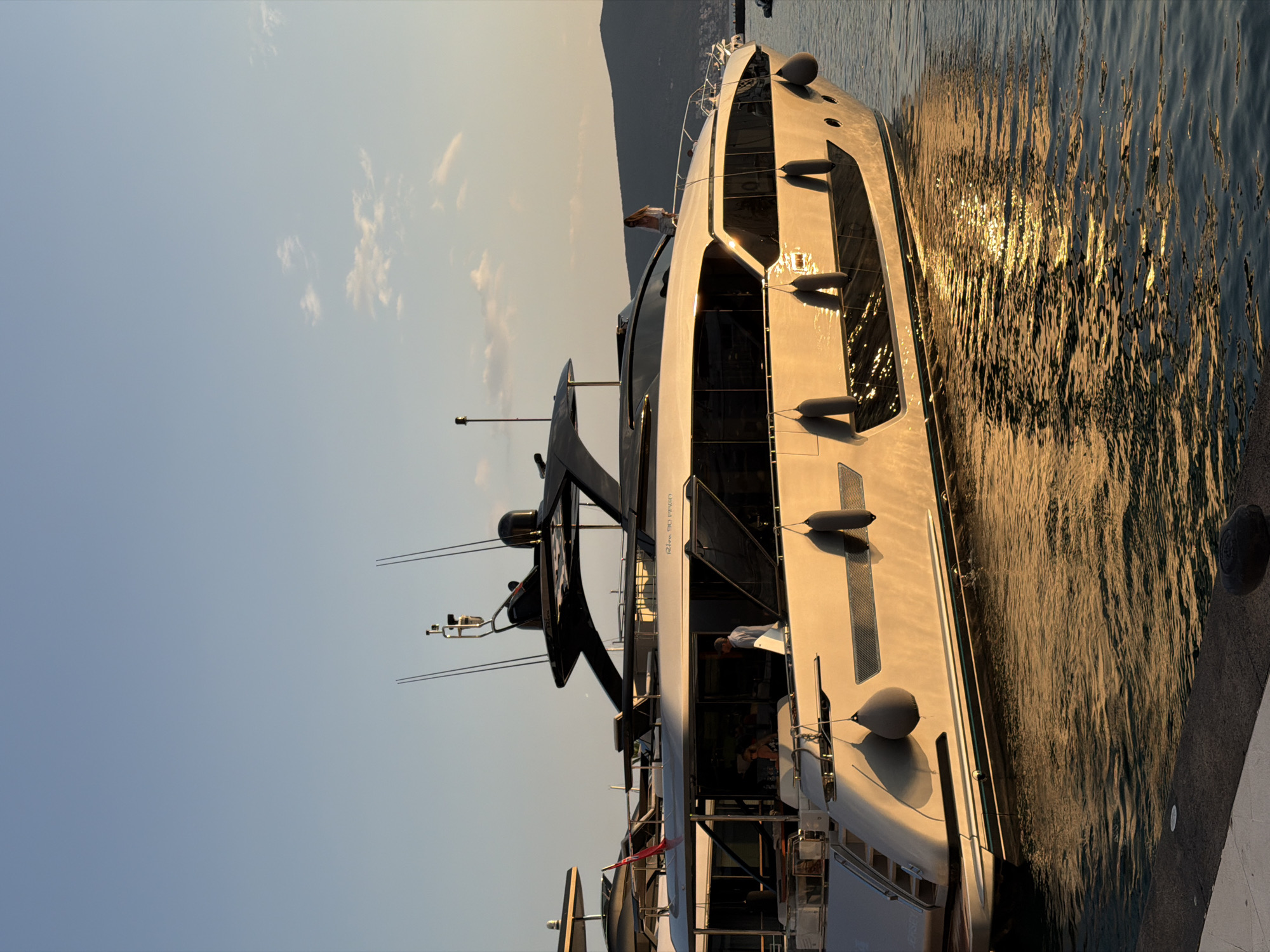 This media file captures a large, modern yacht docked in a harbor, likely in Tivat, Montenegro, during the late afternoon or early evening. The sun is low in the sky, casting a warm, golden light that reflects off the water and the yacht's hull, creating shimmering patterns on the surface.
The primary subject is the sleek, white yacht, which appears to be a luxury vessel with multiple decks and large tinted windows. Several dark, cylindrical fenders are visible along its side, indicating it is moored. A dark, angular superstructure rises from the yacht's stern. There are at least two people visible on the yacht's deck, one standing near an open doorway and another partially visible inside. The activity appears to be a calm, leisurely moment as the day winds down.
In the background, a hazy, mountainous landscape is visible under a clear, pale blue sky with a few wispy clouds. The water is calm with gentle ripples. The overall mood is serene and tranquil, amplified by the warm lighting and the peaceful setting of a harbor. The image conveys a sense of luxury and relaxation. No specific text is clearly legible on the yacht or in the surrounding environment.