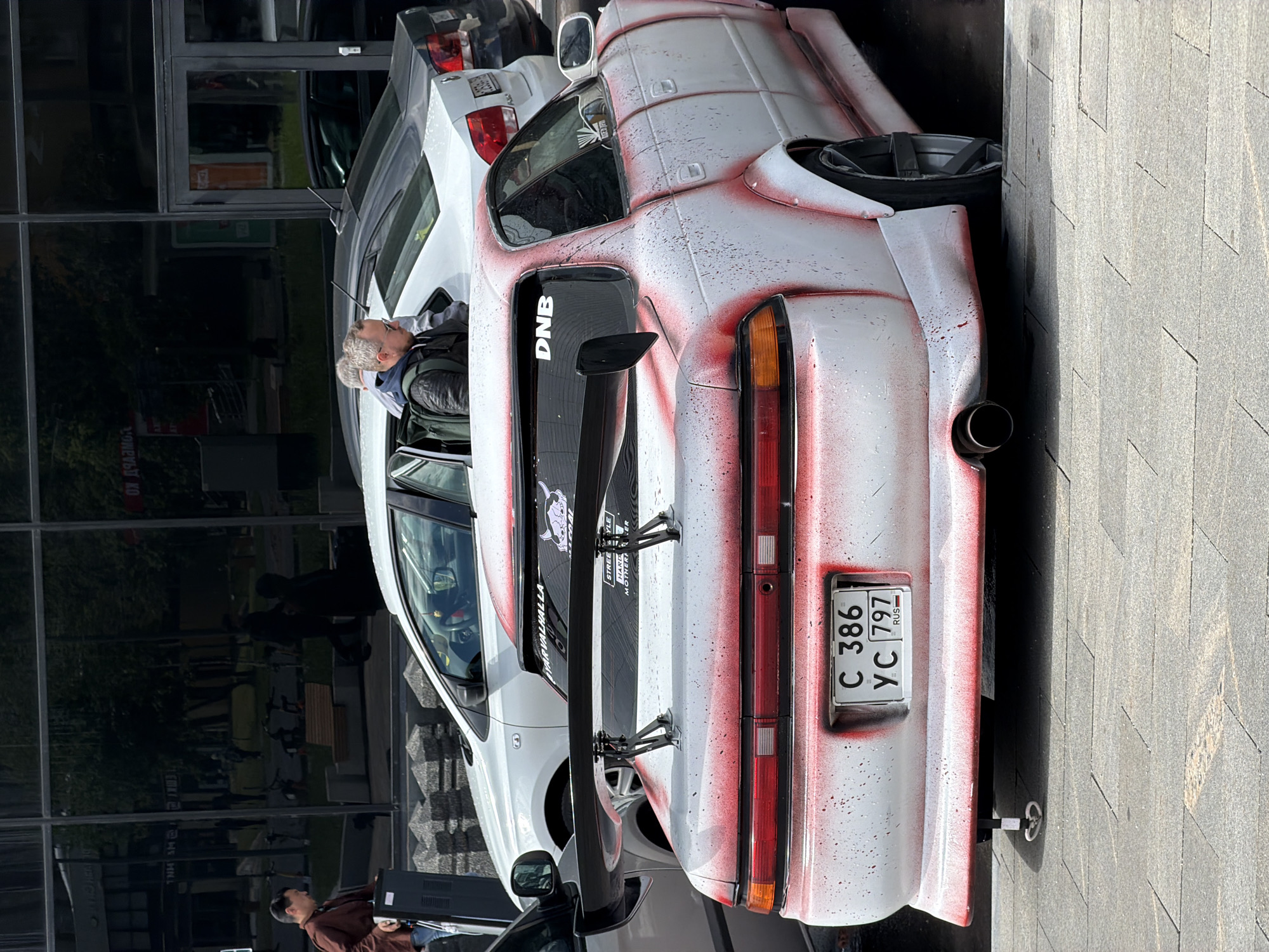A car with Russian license plate C 386 YC 797 is parked on a paved surface in an urban environment. The car is white with red splatter paint on its rear. A man is leaning into the driver's side window, engaging with the car's interior. Behind the first car, another white car is partially visible. The scene is set against the backdrop of a building with reflective glass windows.