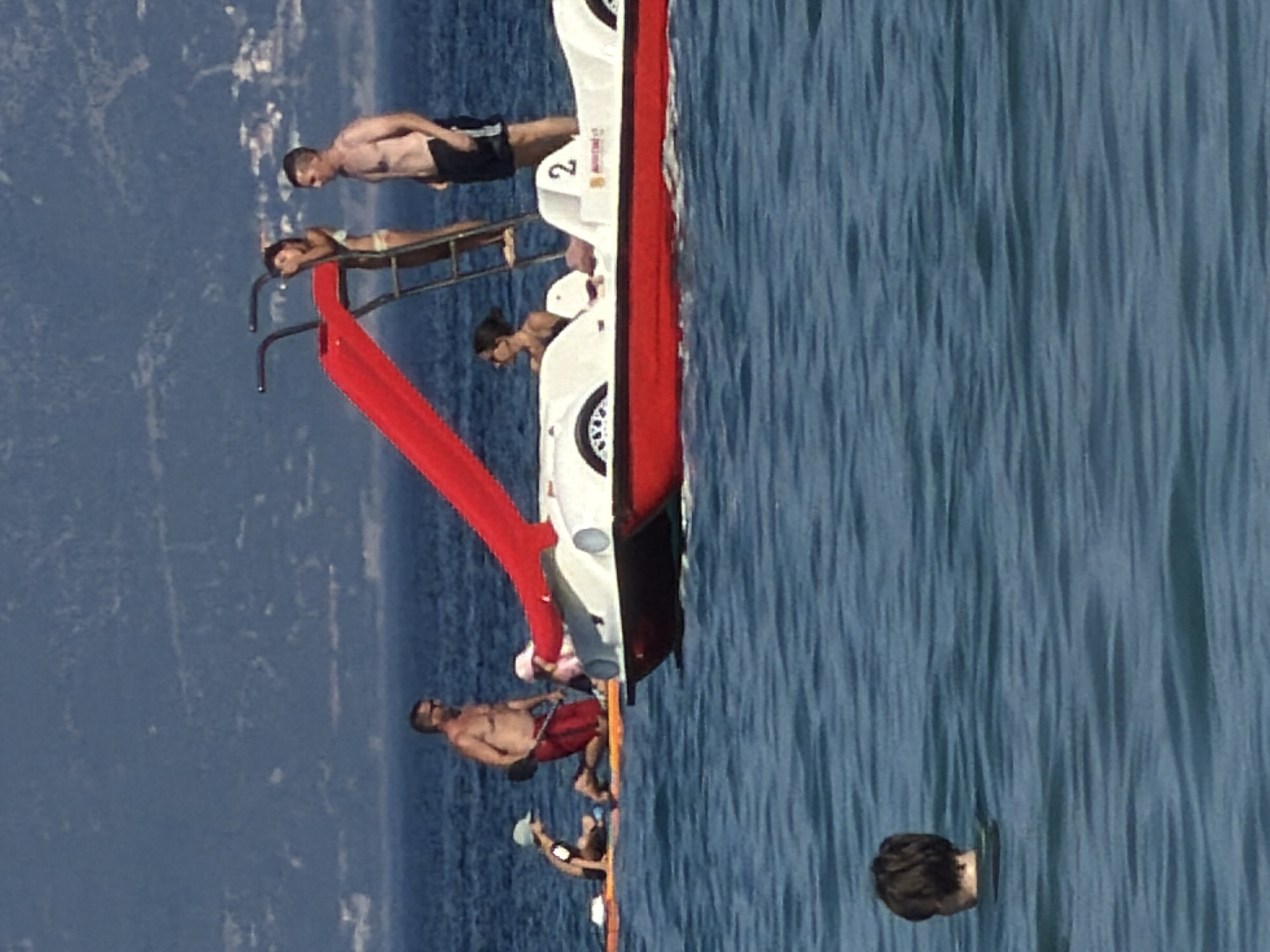 This media file shows several people on a white and red watercraft on the sea. The watercraft appears to be a pedal boat or a small boat with a slide. The scene is set on a bright, sunny day, likely in the afternoon given the lighting. The water is a vibrant blue, indicating clear weather.
The subjects include a man lying on the front of the boat, appearing to sunbathe. Another man is standing on the boat's deck, looking towards the water. A person is visible on the slide, appearing to be about to go down or having just come down. Further back, another man is seated and holding what appears to be a paddle. At the very bottom right, the head of another person is visible, seemingly floating in the water.
The watercraft has a red slide extending from it, inviting people to slide into the water. There are seats on the boat, and some people are dressed in swimwear. One man is wearing red shorts, another black shorts. There's a number "2" visible on the side of the boat. The background shows a hazy, mountainous coastline with scattered buildings, suggestive of a coastal town in a Mediterranean region like Croatia, specifically Općina Baška. The overall mood appears relaxed and leisurely, consistent with a day of fun on the water.