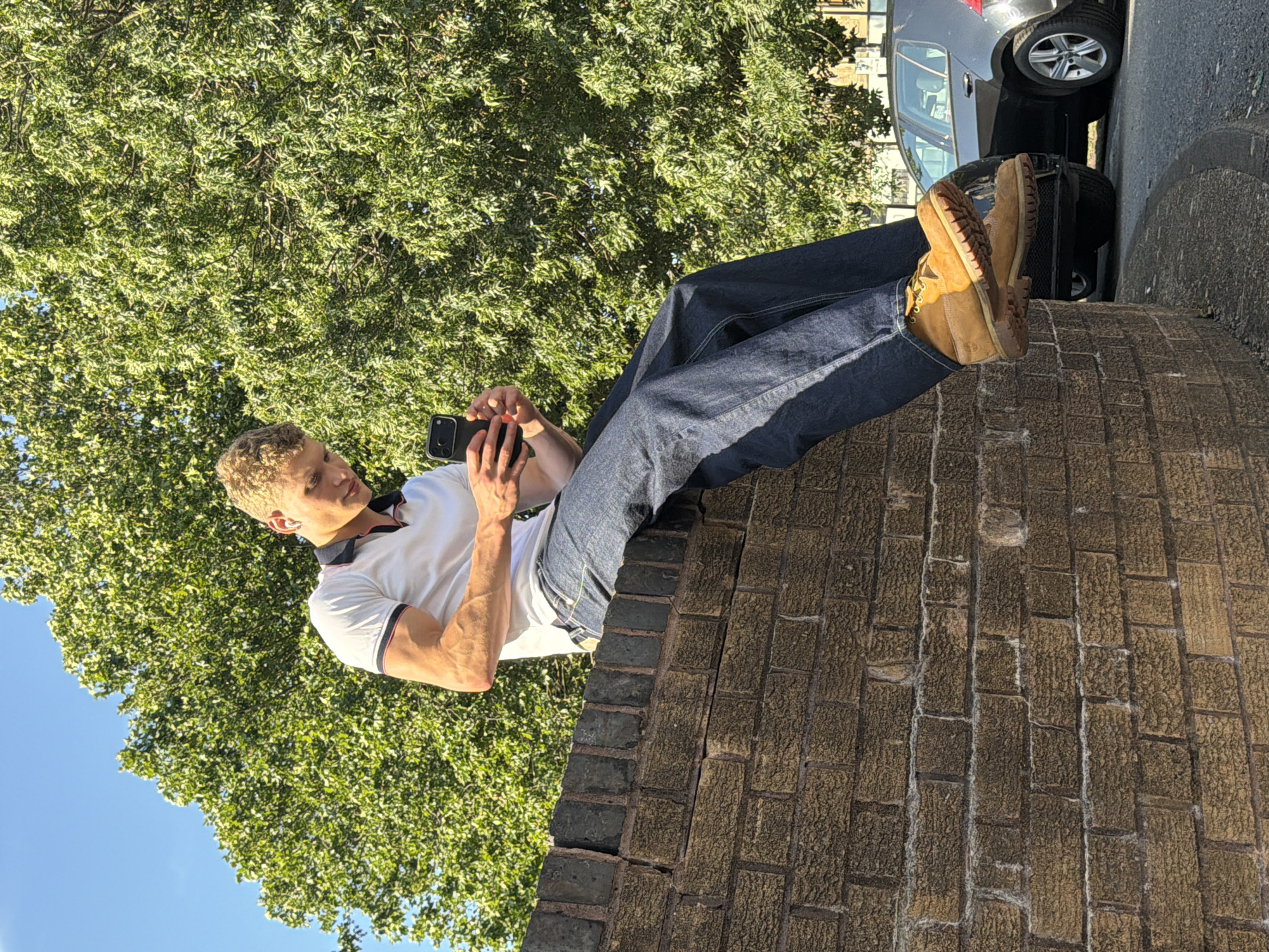 A young man with blonde curly hair is sitting on a textured brick wall, looking at his phone. He is wearing a white polo shirt with blue and red trim, dark wash denim jeans, and tan work boots. The man has earbuds in his ears. The background features a large, leafy green tree and a black car parked on the street. The sky is clear and blue. The scene appears to be outdoors in a residential or urban area.
