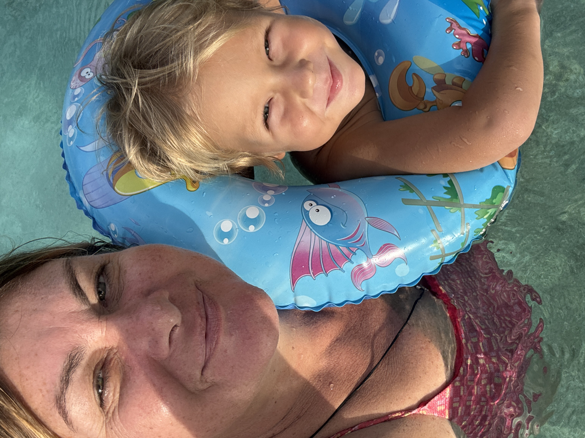 In this close-up, high-angle shot taken in the shallow, clear waters of the ocean near Baan Bang Rak, Thailand, a woman and a young child are positioned together. The woman, on the left, is looking directly at the camera, her face framed by the sunlight. She has light brown hair and appears to be wearing a pink and patterned swimsuit. To her right, the child is also looking towards the camera, with blonde hair, and is wearing a blue inflatable swim vest featuring cartoon sea creatures. The child's arm is visible, and the vest also displays cartoon octopus and coral designs. The water's surface creates ripples and distortions around their submerged bodies and clothing.