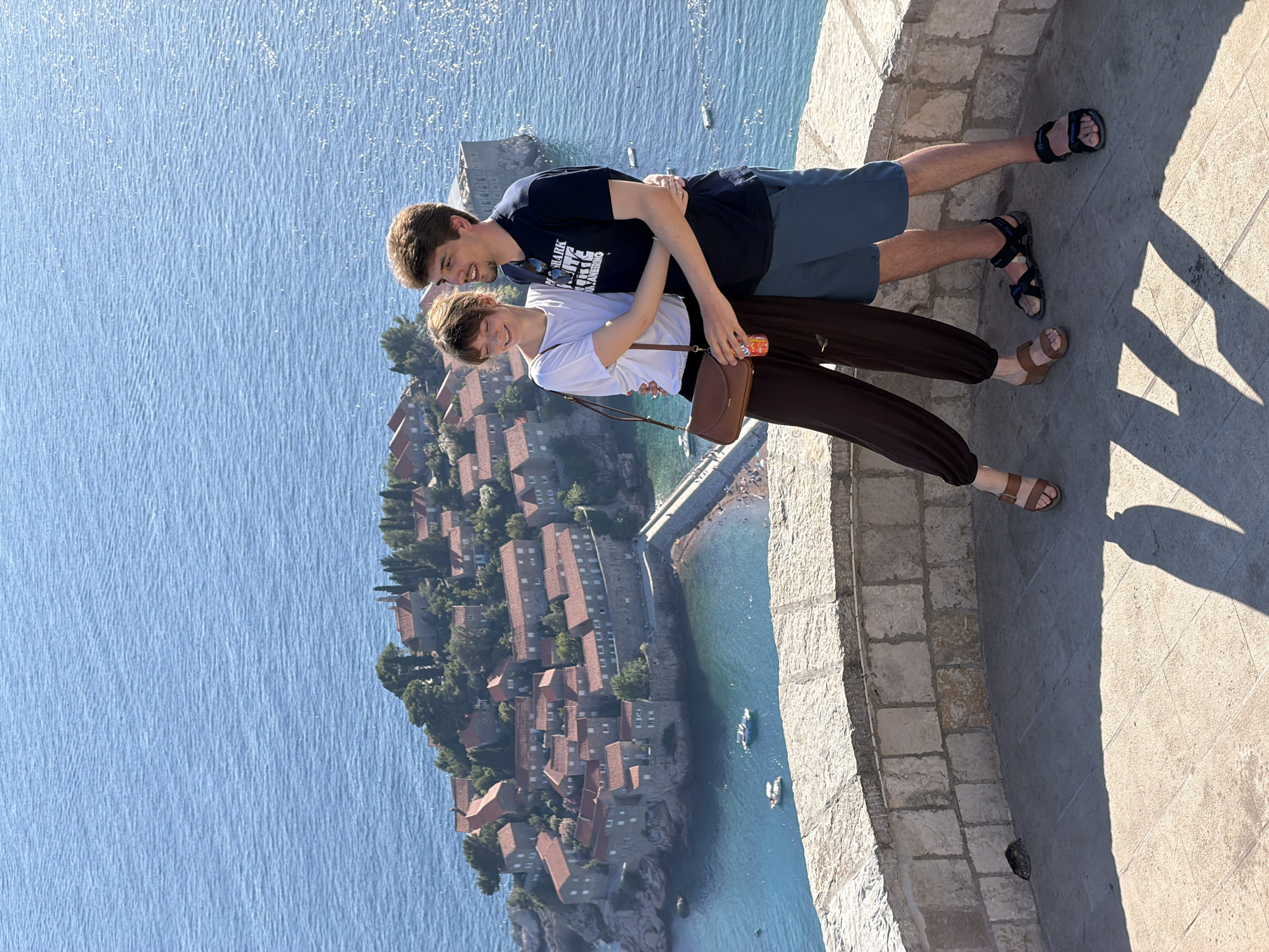 A young couple, a man and a woman, are embracing while standing on a stone-tiled ledge overlooking the Adriatic Sea and the island town of Sveti Stefan in Montenegro. The man, on the right, wears a dark blue t-shirt and blue shorts, with black sandals. He has his right arm around the woman's waist and his left arm around her shoulders. The woman, on the left, is wearing a white t-shirt, dark brown wide-leg pants, and brown sandals. She has her left arm around the man's neck and her right hand resting on his chest. She is holding a can in her right hand. Both are smiling and looking towards the island. The island of Sveti Stefan, connected to the mainland by a narrow causeway, is visible in the background, characterized by its red-tiled roofs and stone buildings, nestled amongst green trees. The sea is a vibrant blue with sunlight glinting on its surface. The ledge they are standing on is constructed of rough stone blocks. The foreground features the shadows of the couple cast on the stone tiles.