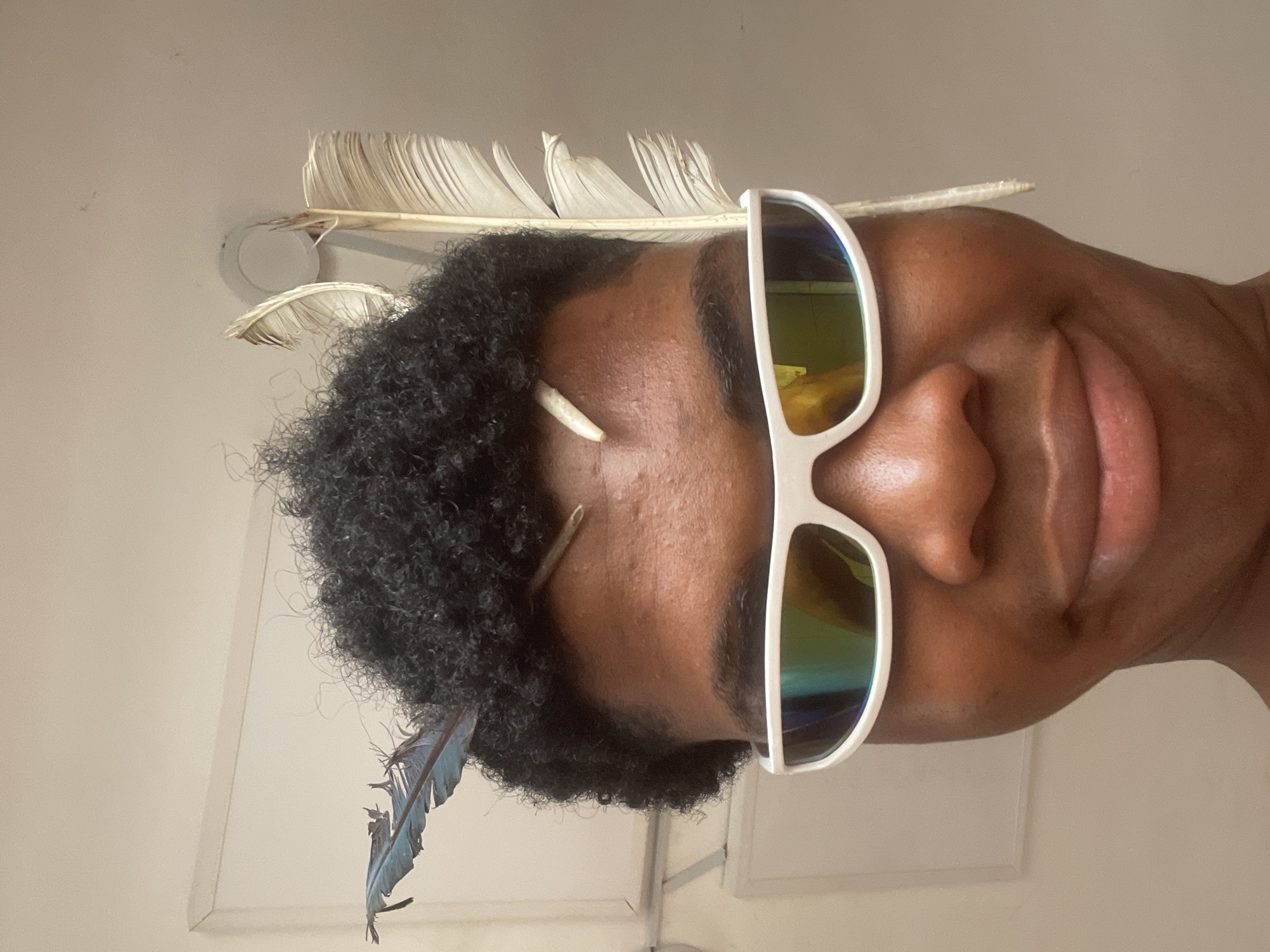 A person with dark, curly hair is looking directly at the camera, wearing white, futuristic-style sunglasses with reflective lenses. Two light-colored feathers are attached to the right side of their head, and a blue feather is attached to the left. A thin, light-colored stick or quill is also protruding from the person's forehead, just above their eyebrows. The background is a plain, light-colored wall with a white rectangular object and a circular light fixture visible. The scene appears to be indoors. The image does not provide any information about the location in the city of Ado, Nigeria.