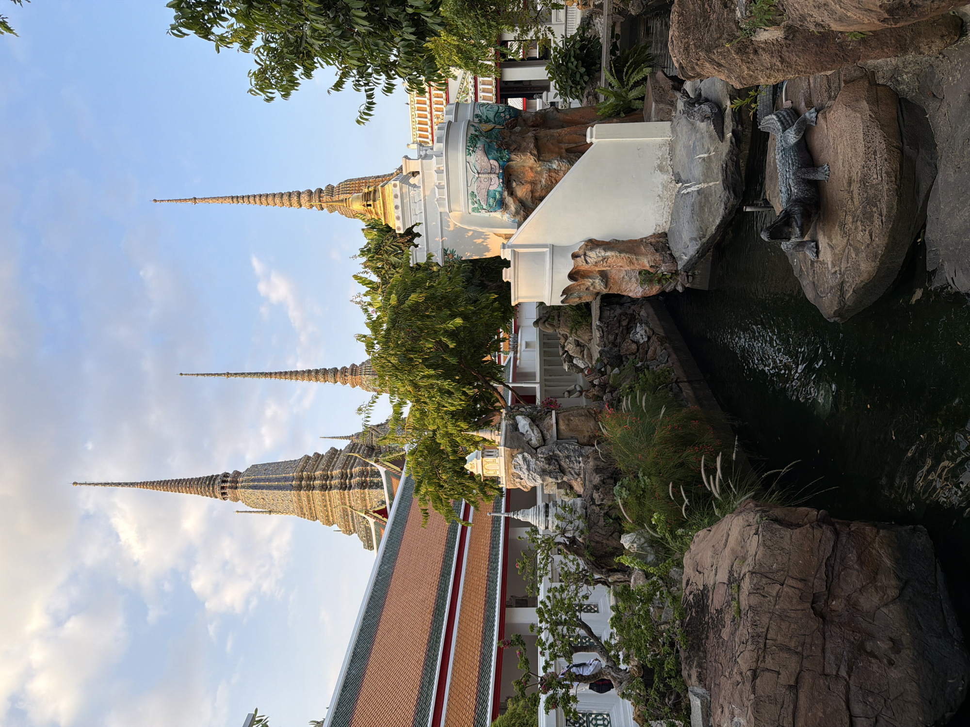 The image captures a scenic view of Wat Pho, a prominent Buddhist temple complex in Bangkok, Thailand. The foreground features a man-made pond or canal with a rocky edge, adorned with lush green vegetation and ornamental plants. A stone sculpture of a crocodile with its mouth open is positioned on a large rock, seemingly emerging from the water. To the right, a white, multi-tiered structure with decorative butterfly motifs rises amidst the rocks. In the background, the iconic spires (chedis) of Wat Pho, known for their intricate details and gold embellishments, pierce the sky, which is a soft blue with scattered clouds. A section of the temple's roof, characterized by orange tiles and green trim, is also visible. The overall scene is bathed in the warm light of what appears to be late afternoon or early morning.