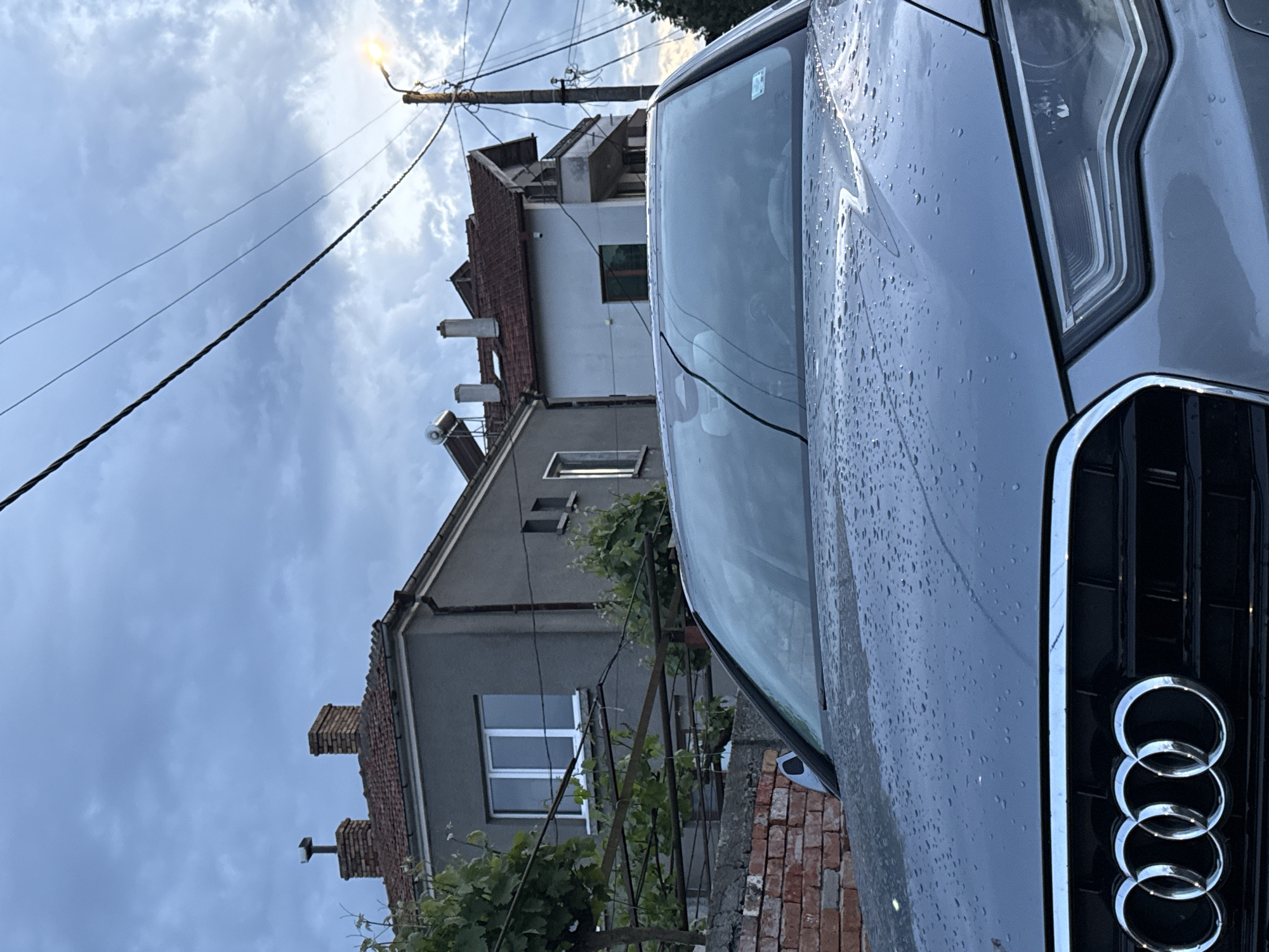 The image shows the front of a gray Audi vehicle parked on a street in Burgas, Bulgaria. The vehicle's windshield and hood are covered in water droplets, suggesting it has recently rained. The Audi logo is prominently displayed on the front grille.
Behind the car, there are two buildings. The closest building to the car is gray, with white-framed windows and a brick chimney. The other building has a red-tiled roof and a light-colored wall. A street lamp is visible in the upper left of the image. Utility wires are strung across the sky.
The sky is overcast with visible clouds. Vegetation is present near the brick wall.