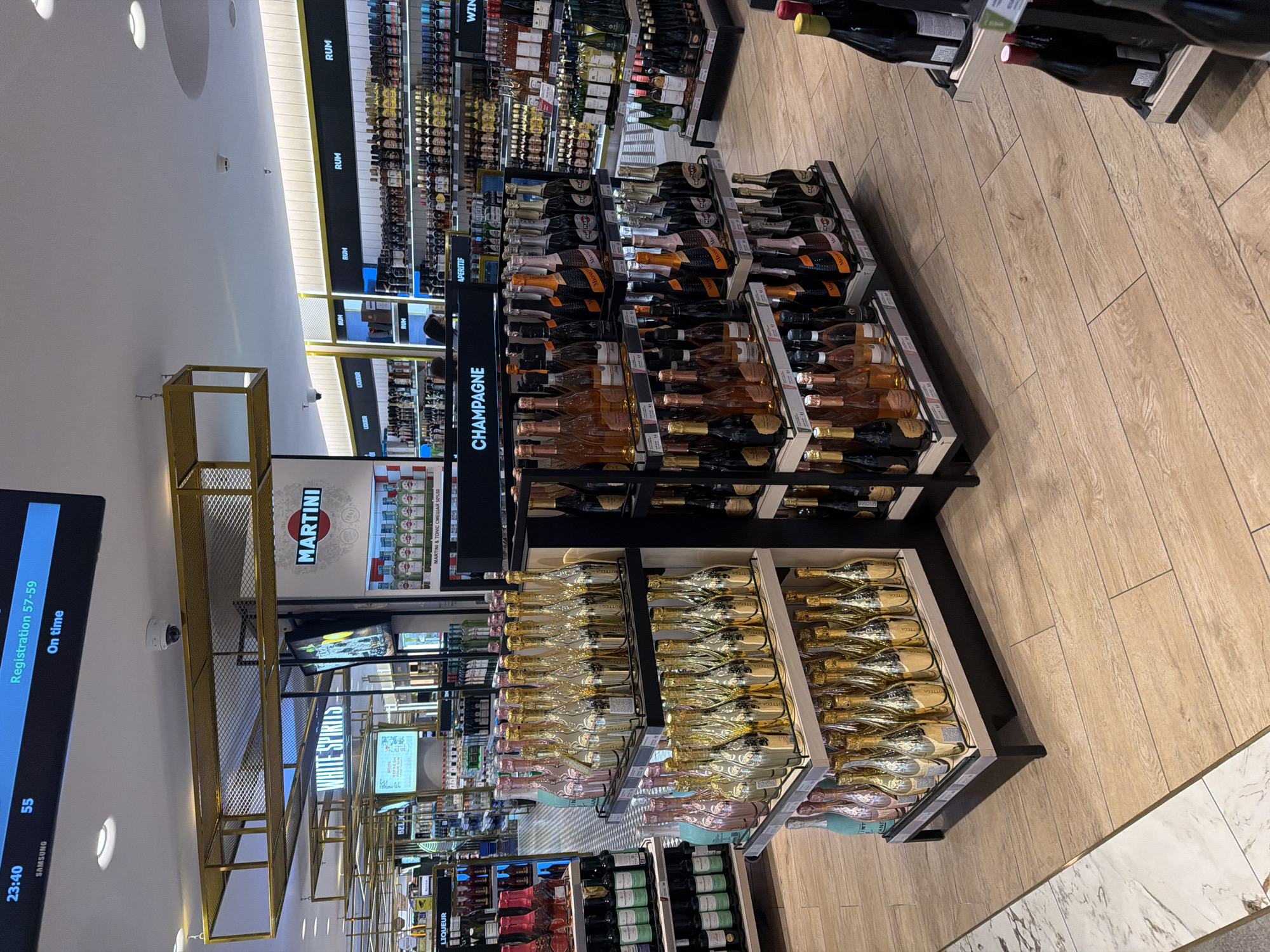The image depicts a duty-free shop, likely at an airport, in Saint Petersburg, Russia, as indicated by the flight information display at the top left of the image showing "Registration 57-59" and "On time" at "23:40". The scene is brightly lit and features shelves stocked with various alcoholic beverages.
In the foreground, on the left, is a display of champagne bottles, with rows of gold-colored and pink bottles on tiered shelves. To the right of the champagne display is another display of wine bottles, some in dark glass and others in lighter glass. Further back, on the right side of the image, is a section of rum bottles, and beyond that, shelves filled with various other spirits and liqueurs, including a prominent "MARTINI" display. The flooring is a light-colored wood-effect tile, with a marble-effect tile border on the far left.
The background shows more rows of merchandise, suggesting a large retail space. There are no discernible people in the immediate foreground, but two individuals are partially visible in the middle ground, browsing the shelves. The environment appears clean and organized, typical of a commercial retail setting. The overall impression is of a well-stocked liquor store, likely catering to travelers.