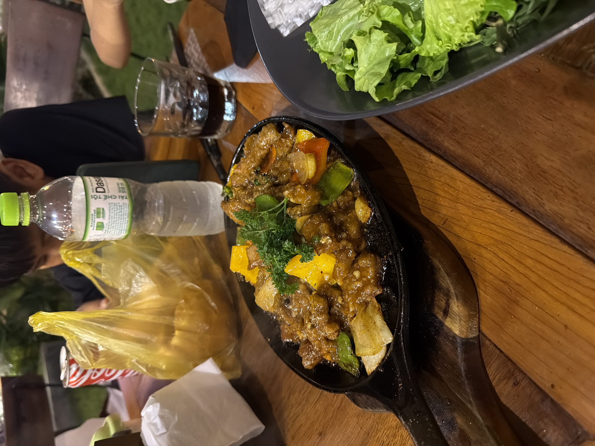 The image depicts a nighttime, outdoor dining scene focused on a wooden table laden with food and beverages. In the foreground, a black cast-iron sizzling plate on a wooden base holds a stir-fried dish consisting of chunks of meat in a dark, glossy sauce, mixed with yellow bell peppers, green bell peppers, and white onion slices. A garnish of fresh green parsley sits atop the dish, from which steam is visibly rising. To the right of the sizzling plate, a black ceramic bowl contains fresh green lettuce leaves and crumpled white rice paper wrappers. On the table further back, a red Coca-Cola can and a clear plastic bottle of Dasani water with a green cap and Vietnamese labeling ("TẢI CHẾ TÔI") are present. A clear glass containing a dark liquid with ice cubes is also visible. Two individuals are partially visible in the blurred background: one person with dark hair and a dark shirt is seated to the left, looking down or away, and a portion of another person's arm is visible on the far right. The table is constructed from light-brown wooden planks. The background shows blurred green foliage and dark structures under artificial illumination, indicating an outdoor or semi-outdoor setting at night. The presence of Vietnamese text on the Dasani bottle confirms the location as Vietnam. However, the specific identification of "Nha Trang Ward" cannot be forensically determined from the visual information within the image.