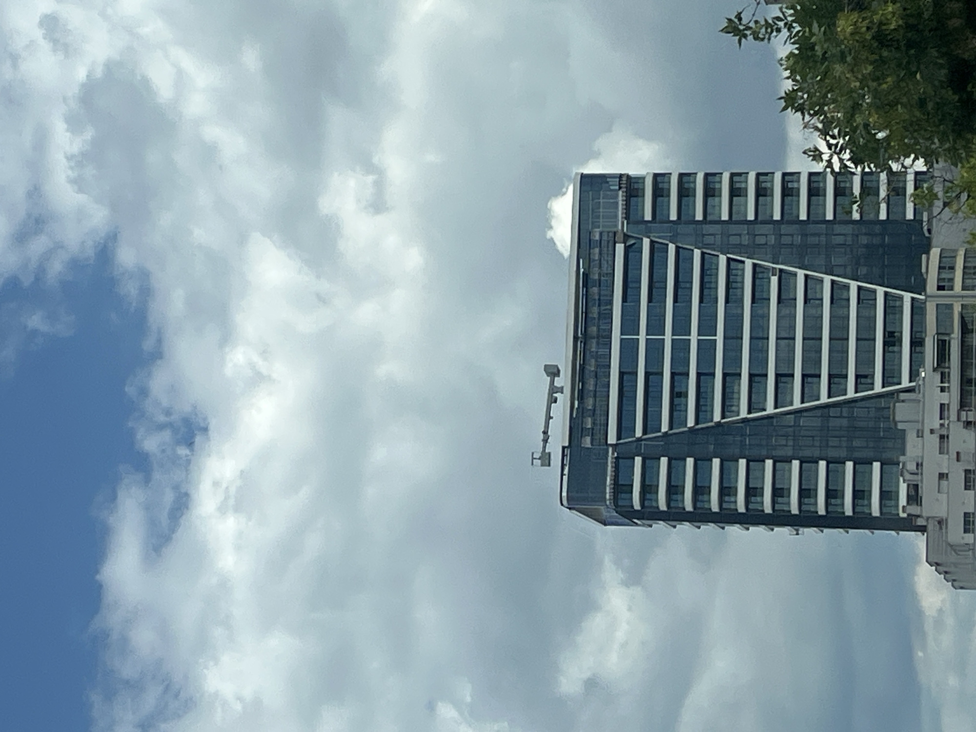 A tall, modern skyscraper with a dark glass facade and white horizontal accents dominates the center of the frame against a cloudy sky. A piece of equipment, possibly a crane or antenna, is visible on the flat roof of the building. In the foreground, the tops of green trees are partially visible on the right side of the image. The lower portion of the frame shows the edge of a lower, lighter-colored building. The scene appears to be in an urban environment.