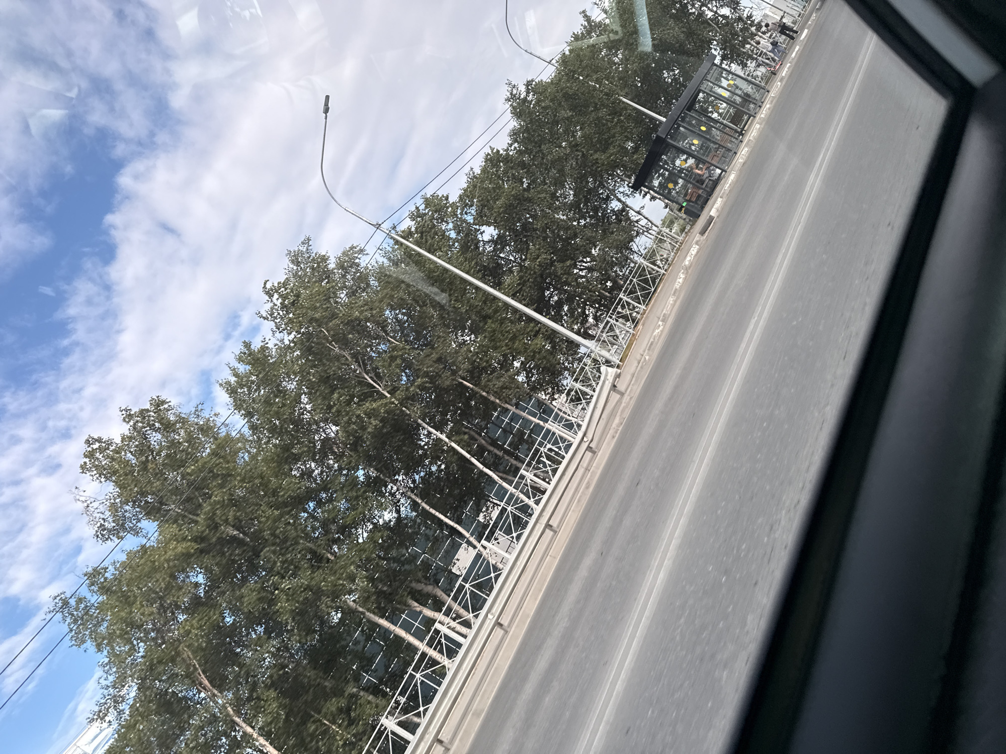 The image shows a view of a street scene in Yuzhno-Sakhalinsk, Russia, taken from inside a vehicle. The perspective is angled, giving a dynamic feel to the scene. The foreground is dominated by the interior of the vehicle, with a dark window frame cutting across the bottom.
The street is paved and appears to be a multi-lane road. A white metal railing runs along the side of the road, bordering a sidewalk or embankment. Behind the railing, a line of trees with full green foliage stands tall, their branches reaching towards the sky. The trees appear to be deciduous, with slender trunks. A modern-looking glass bus stop shelter is visible on the right side of the image, with yellow circular motifs on its exterior. Some people are visible near the bus stop, one standing and two or three appearing to be seated inside.
In the background, the sky is a mixture of blue sky and white, fluffy clouds. A street light with a dark fixture is visible on a curved pole above the trees. Electrical wires are strung across the upper portion of the image. The overall impression is of an urban environment on a clear day, with a focus on the natural elements of trees and sky juxtaposed with man-made infrastructure like the road, railing, and bus stop.