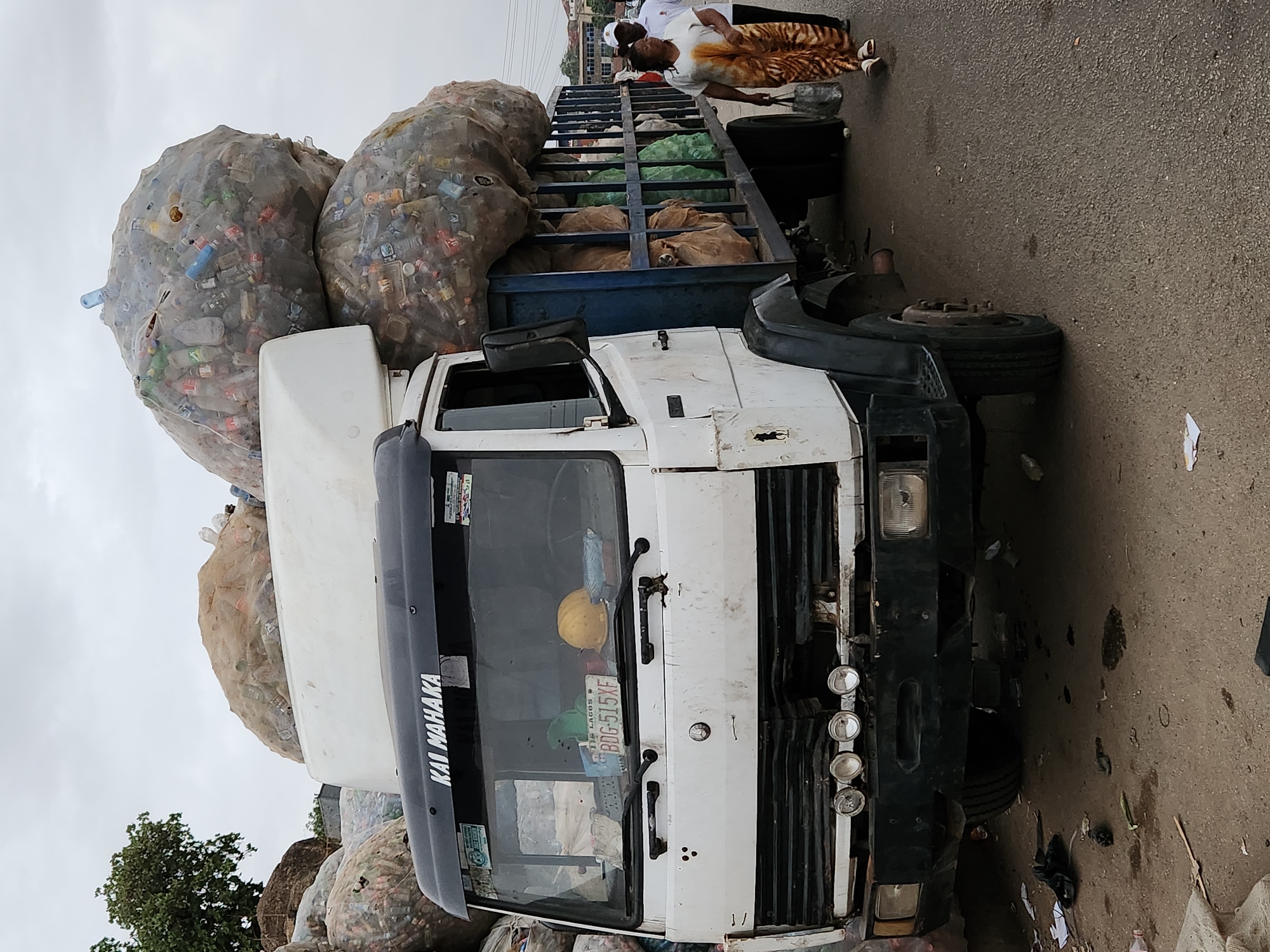 The image depicts a white truck heavily loaded with large, netted bags filled with plastic bottles. The truck is on a paved surface, likely a street. A person wearing a white top and patterned brown skirt is standing next to the truck's cargo bed, holding a tool, possibly a shovel. The truck has "KAI MAHAKA" printed on the front. The license plate visible on the truck reads "BDG-515XF". Based on the name "KAI MAHAKA," which is a Lagos State Waste Management Authority vehicle, it's likely that the truck is involved in waste collection and the location is Kabong, Nigeria. The background shows a cloudy sky and hints of buildings and greenery. The surrounding street area is littered with debris.