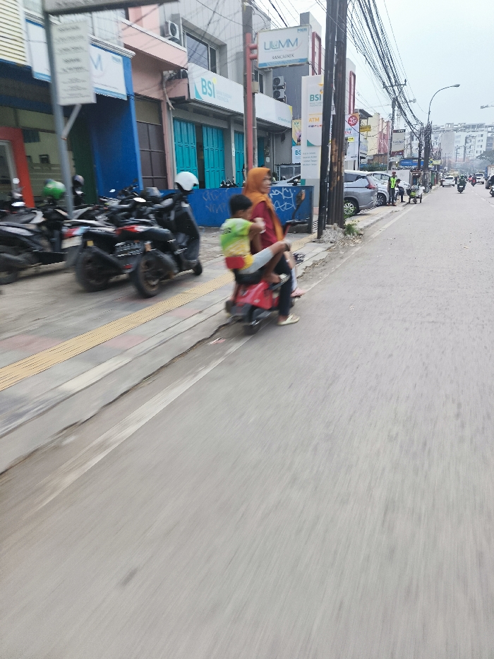 A woman wearing an orange headscarf is riding a red moped with a small child in the front, facing forward, along a street in Jatinangor, Indonesia. The moped is stopped near a sidewalk. A row of parked cars is to the woman’s left, facing the opposite direction. There are other vehicles, such as a car and motorbikes, parked on the street. Buildings are in the background. The road appears to be paved, with a sidewalk on the side. The sky is partly cloudy. The street is mostly empty. The woman is wearing a red shirt and the child is wearing a yellow shirt and green backpack. They are facing forward. The moped has red handlebars. The woman’s right foot is on the moped’s footrest, and her left foot is on the ground. The child’s legs are around the woman’s waist. The image was taken during the day.