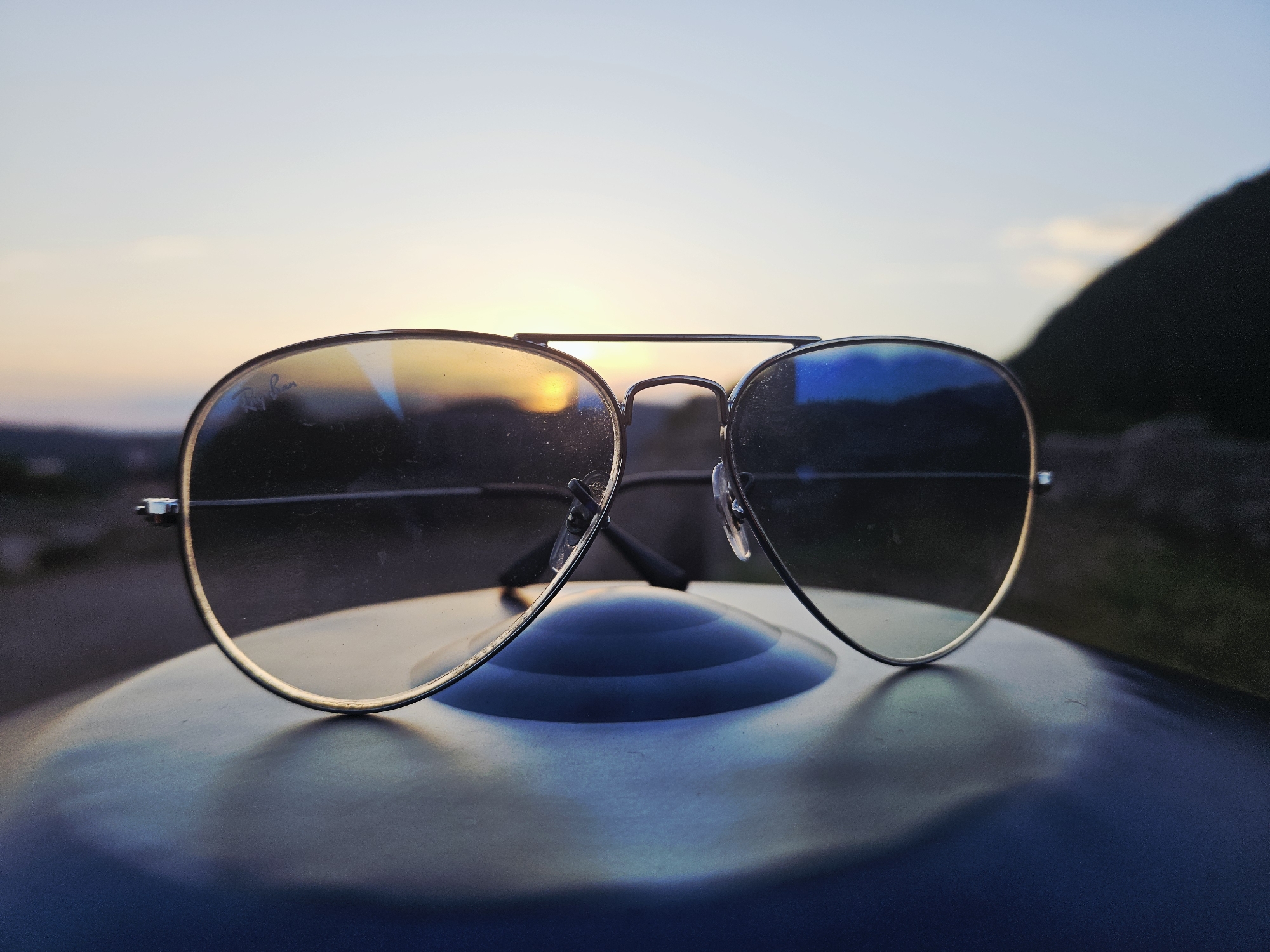 The image displays a close-up of a pair of silver-framed aviator-style sunglasses resting on a dark blue, curved surface. The sunglasses are prominently in the foreground and sharp focus.
The scene captures either a sunrise or sunset, with the horizon featuring a warm, golden glow from the sun. This light is strongly reflected on the left lens of the sunglasses, while the right lens reflects cooler, bluer hues from the sky. The sky above the horizon is a gradient of light blue, transitioning to a deeper blue further up. In the background, out of focus, are faint outlines of distant hills or mountains, suggesting an outdoor setting.
The "Ray-Ban" logo is subtly visible etched on the upper left portion of the left lens. The surface beneath the sunglasses appears smooth and slightly reflective, possibly part of a vehicle like a car dashboard or hood. The overall mood conveyed is one of peace and serenity, characteristic of dawn or dusk. No activity is depicted; it is a still life photograph.