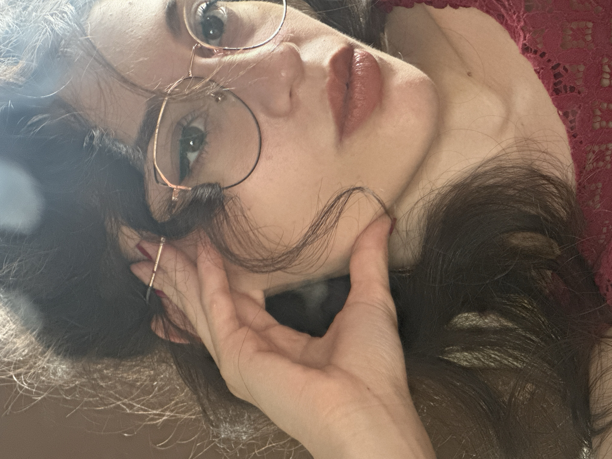 A close-up, indoor, waist-up shot captures a young woman with long, dark, wavy hair. She is wearing rose gold, hexagonal-shaped eyeglasses and a dark red lace top. Her right hand is raised to her face, with her index finger touching her temple and her thumb resting on her cheekbone. Her lips are painted with a matte, reddish-brown lipstick. Her eyes are open and looking towards the camera, with subtle winged eyeliner. The lighting suggests an indoor setting with light casting shadows on her hair and face. The background is a muted, textured brown, possibly a wall. There is no information to determine the location in Sofia, Bulgaria.