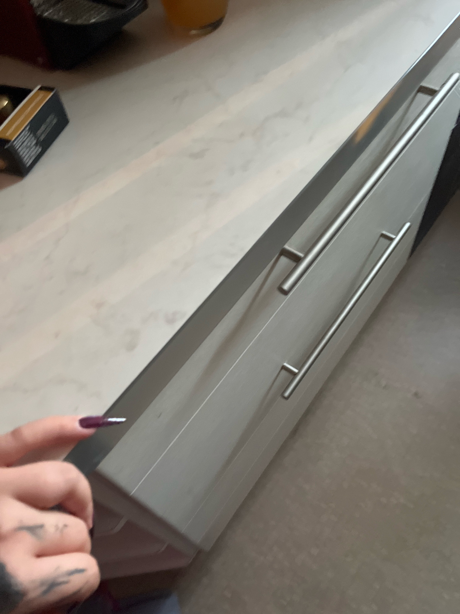 The image shows a close-up view of a kitchen counter with two drawers. A hand with dark purple nail polish is visible in the foreground on the left, with its index finger pointing towards the drawers. The counter is made of a light-colored material with a subtle marble-like pattern. The drawers have sleek, silver bar handles. To the left of the counter, a small black box with a yellow stripe is visible, possibly containing matches. The background is out of focus, but appears to be a kitchen environment. There is no identifiable information in the image to suggest a location in Moscow, Russia.