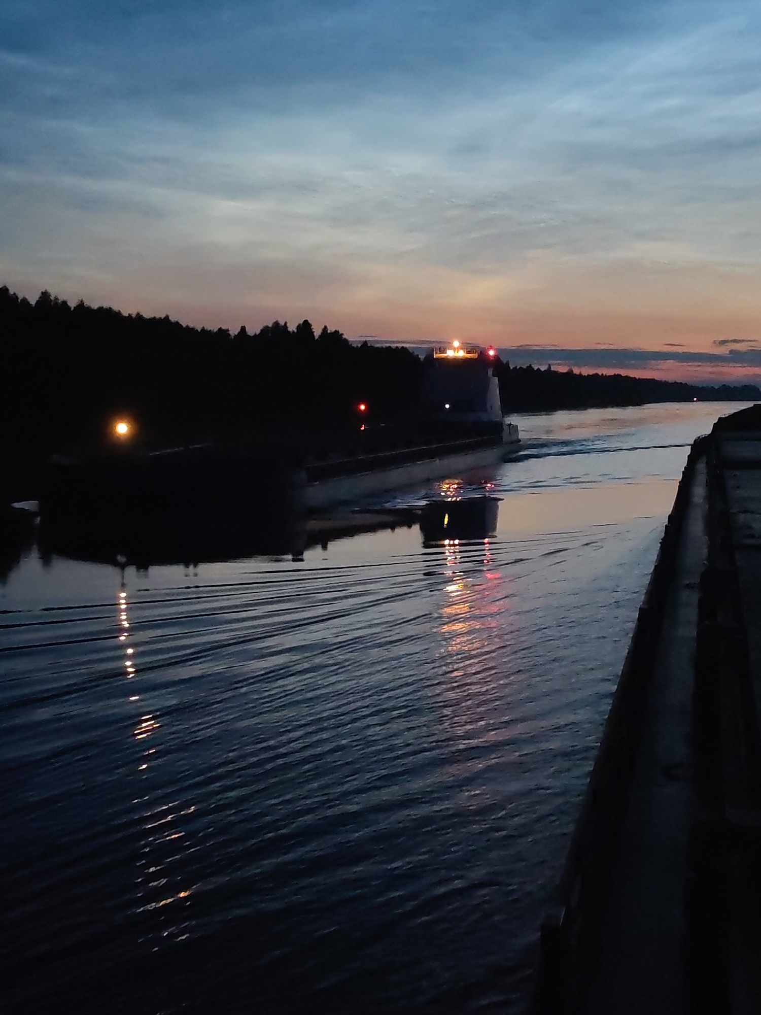 The image depicts a twilight scene on the Svir River near Vytegra, Russia, likely within or near a lock system. A large, dark barge or vessel is visible in the middle of the waterway, illuminated by its own lights, which cast reflections on the rippling water. The barge appears to be moving or stationary within a canalized section of the river, indicated by the concrete walls on either side. The sky is a gradient of deep blue at the top, transitioning to lighter hues of pink and orange near the horizon, characteristic of sunset or sunrise. Silhouetted trees line the banks of the river, creating a dark and dense forest. The overall atmosphere is serene and somewhat industrial, given the presence of the barge and the lock. There are no discernible people in the image.