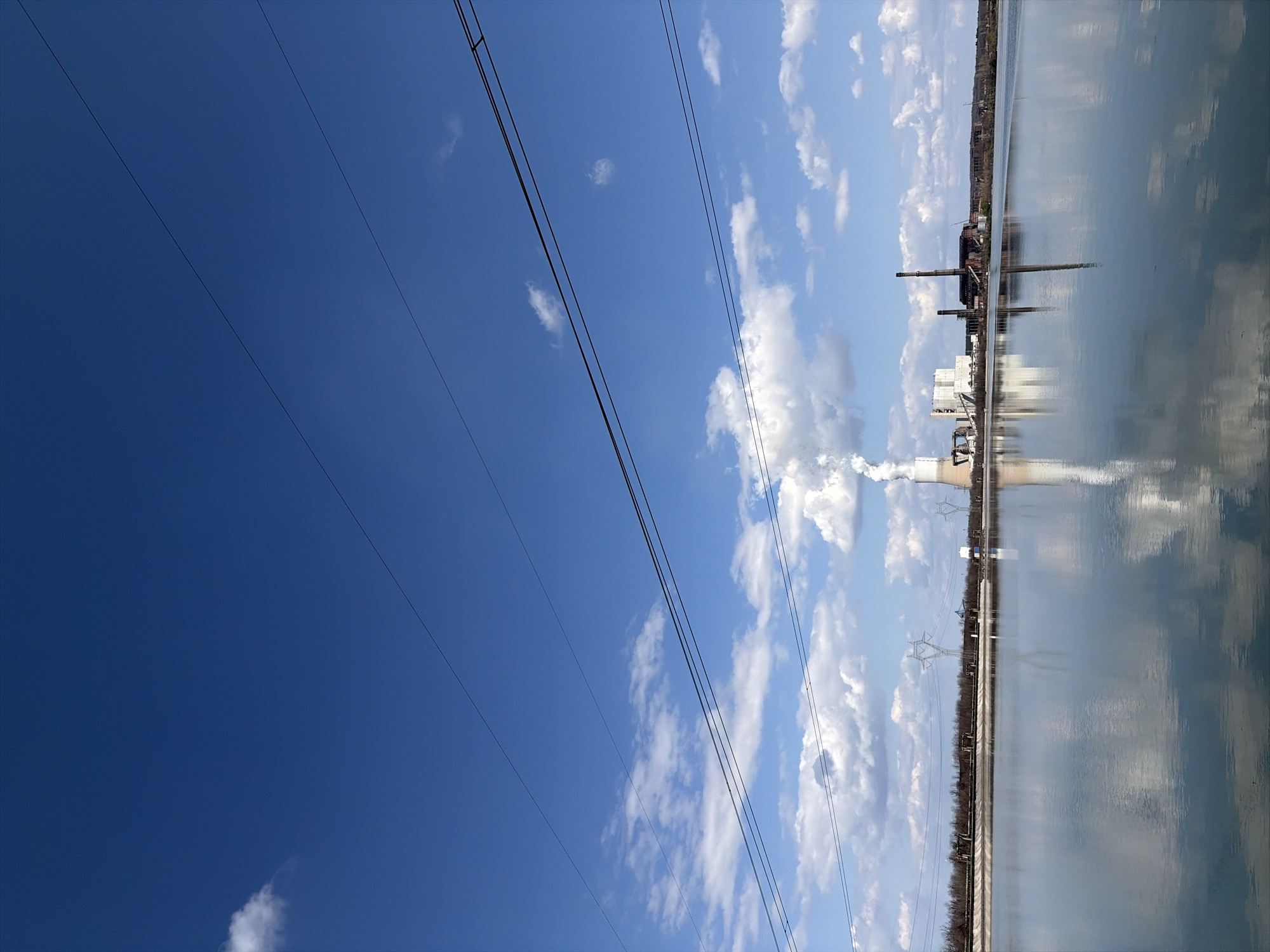The image shows a horizontal view of the Maritza East 1 Thermal Power Plant, located near Galabovo, Bulgaria, reflected in a body of water. The sky is blue with scattered cumulus clouds. Overhead power lines stretch across the upper left portion of the image. The power plant includes smokestacks and cooling towers, with steam rising from one of the towers. A strip of vegetation is visible on the far side of the water.