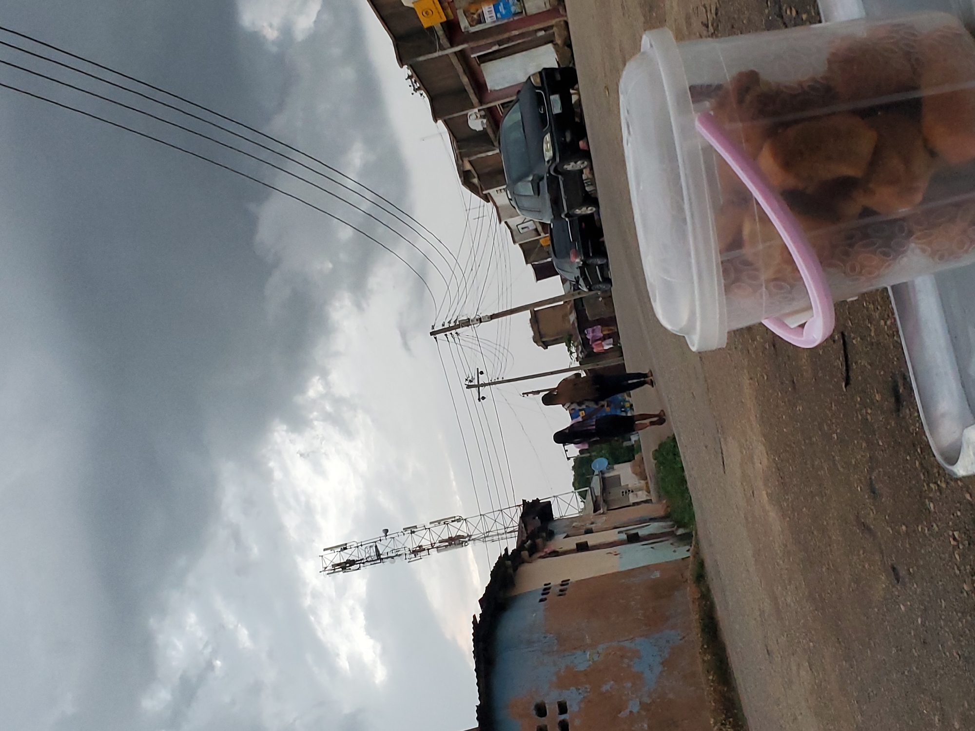 The image shows a street scene in Jos, Nigeria, under a cloudy sky, suggesting it might be overcast or approaching rain. The photograph is taken from a low angle, looking upwards and to the side, with a plastic container filled with food items in the foreground on the right. The container appears to be clear plastic with a pink-trimmed lid.
In the background, there are several structures, including buildings that appear to be shops or homes, and utility poles with numerous wires stretching across the frame. A few vehicles are visible, including what looks like a black SUV parked in front of a building. A telecommunications tower stands tall in the mid-ground on the left side of the image.
A few people are seen walking on the street, their backs turned to the camera. They are dressed casually. The overall atmosphere appears to be that of a typical day in a bustling urban or semi-urban environment. There is no visible text in the image. The lighting suggests it could be daytime, possibly late afternoon given the overcast conditions.