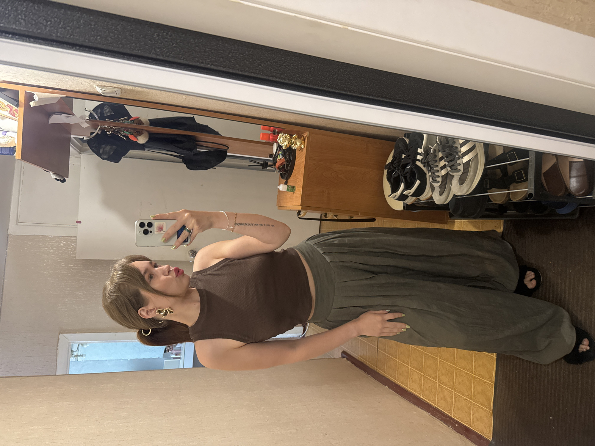 A young woman with light brown hair pulled into a ponytail, wearing a brown tank top, olive green wide-leg pants, and black flip-flops, stands in a hallway. She is taking a selfie in a full-length mirror. She has large gold earrings and a thin bracelet on her right arm. Her left arm has a tattoo with text. The hallway has tiled yellow flooring and beige walls. To the right of the woman, a wooden dresser is visible, with items on top including a small golden dog figurine and a bottle. A shoe rack with several pairs of shoes is also present. The background also shows a door and a reflection of a room with a shelf holding various items. The scene appears to be set in a residential apartment in Chaikovsky, Russia.