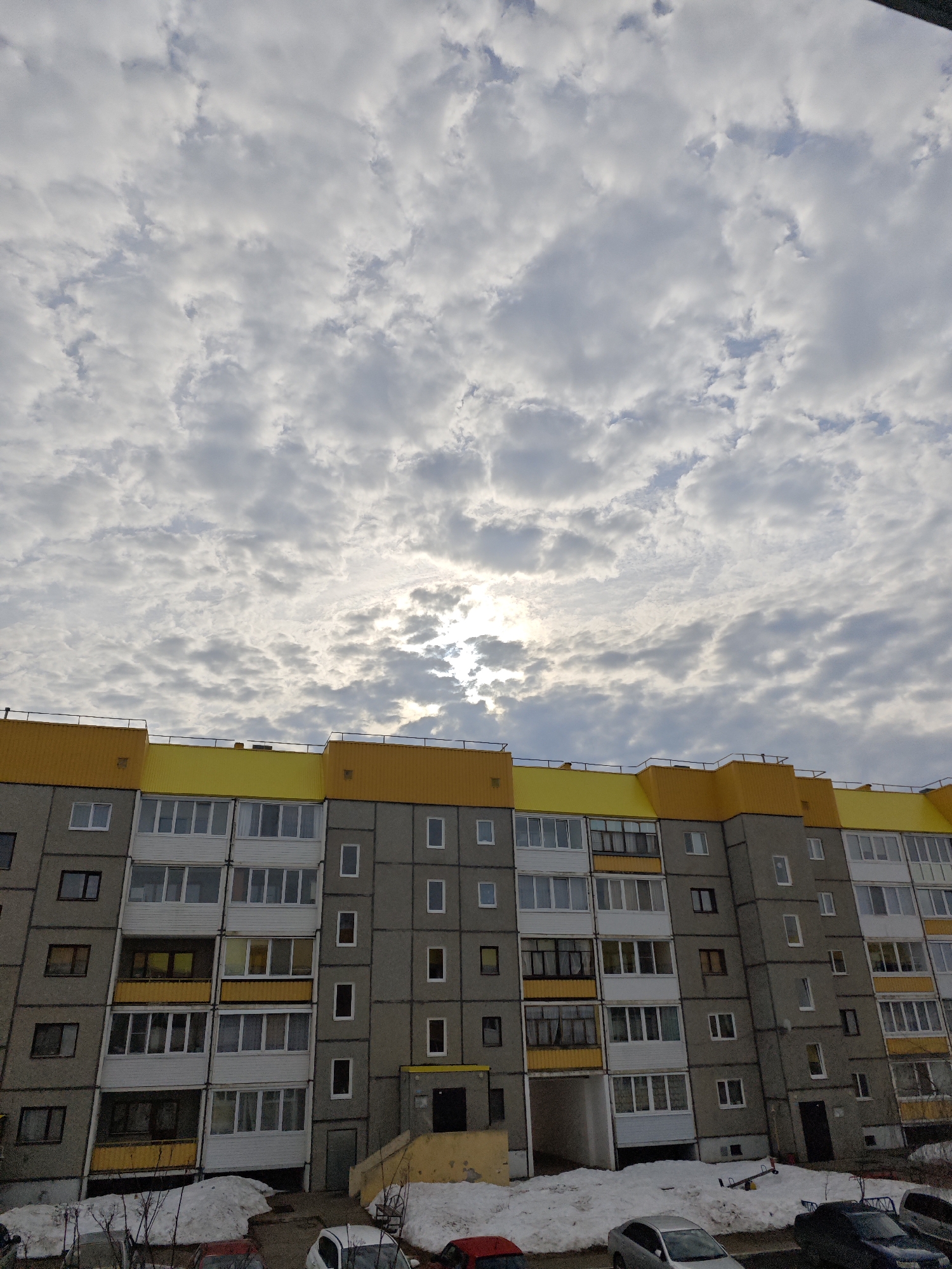 The image depicts a five-story residential building in Марковский, Russia, under a sky filled with stratocumulus clouds, partially obscuring the sun. The building has a concrete facade with a yellow roofline. The windows are a mix of white and brown frames, and some balconies are visible. The ground is partially covered with snow, and several cars are parked in front of the building. There are no people visible in the frame.