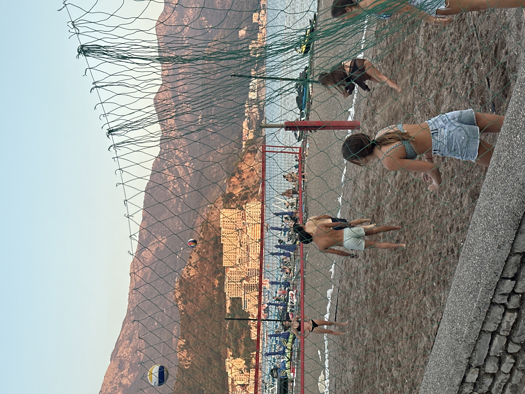 In Boreti, Montenegro, a group of young people are playing beach volleyball at sunset. The scene is set on a sandy beach with the Adriatic Sea and a hillside town in the background. A green net divides the volleyball court, with a volleyball suspended in the air. Several individuals are actively engaged in the game, some in mid-air, while others stand ready to receive or serve. Beach umbrellas and lounge chairs are visible near the water's edge, suggesting a lively tourist area. The overall atmosphere is one of leisure and recreation.
