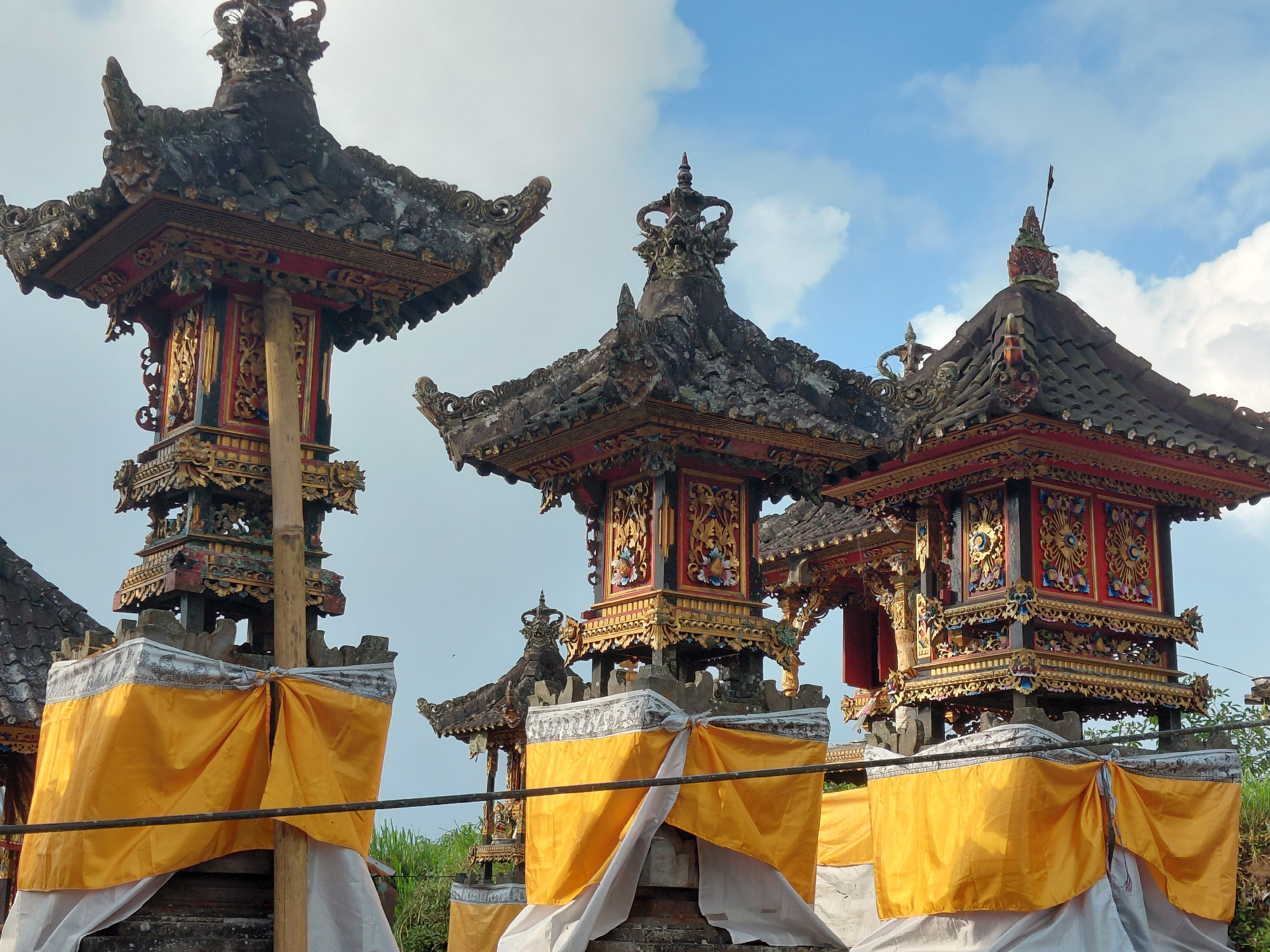 The image shows a close-up view of several intricately decorated Balinese temple structures, specifically focusing on the tiered roofs and ornate carvings. The structures are adorned with gold and red paint, and feature detailed patterns and figures. Yellow and white fabrics are draped around the lower sections of the structures, likely for decoration or ritualistic purposes. In the background, the sky is visible with scattered white clouds. The image appears to have been taken outdoors in the city of Sukasada, Indonesia, judging by the architectural style. No people are visible in the image.