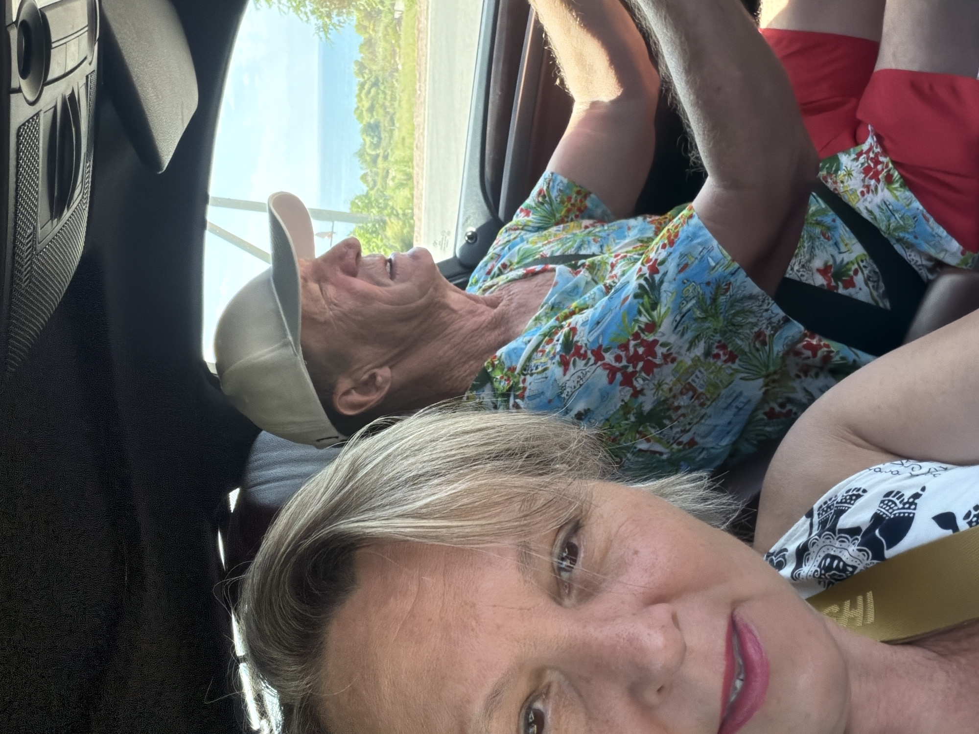In the city of Unknown, Unknown, two people are in a car on a sunny day. A woman in the foreground, with blonde hair and pink lipstick, is looking towards the camera. Next to her, a man wearing a light-colored cap and a colorful shirt is driving. He has his right arm on the steering wheel. The background shows a view of the ocean and greenery through the car window. The scene appears to be a candid shot taken from inside the vehicle.