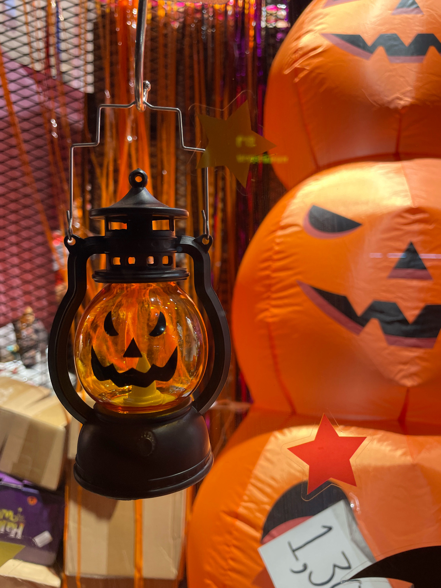 A close-up, vertical, eye-level shot shows a Halloween-themed display, possibly in a shop in Pattaya City, Thailand. In the foreground, a small, black lantern with an orange, pumpkin-shaped glass globe hangs from a metal stand. The glass globe features a carved jack-o'-lantern face. Behind the lantern, a stack of large, inflatable orange pumpkins with carved black jack-o'-lantern faces are visible. One pumpkin has a red star sticker on it. In the lower left portion of the image, cardboard boxes are stacked, some with purple Halloween-themed packaging. The background is blurred and appears to be a display of orange and yellow streamers or lights.