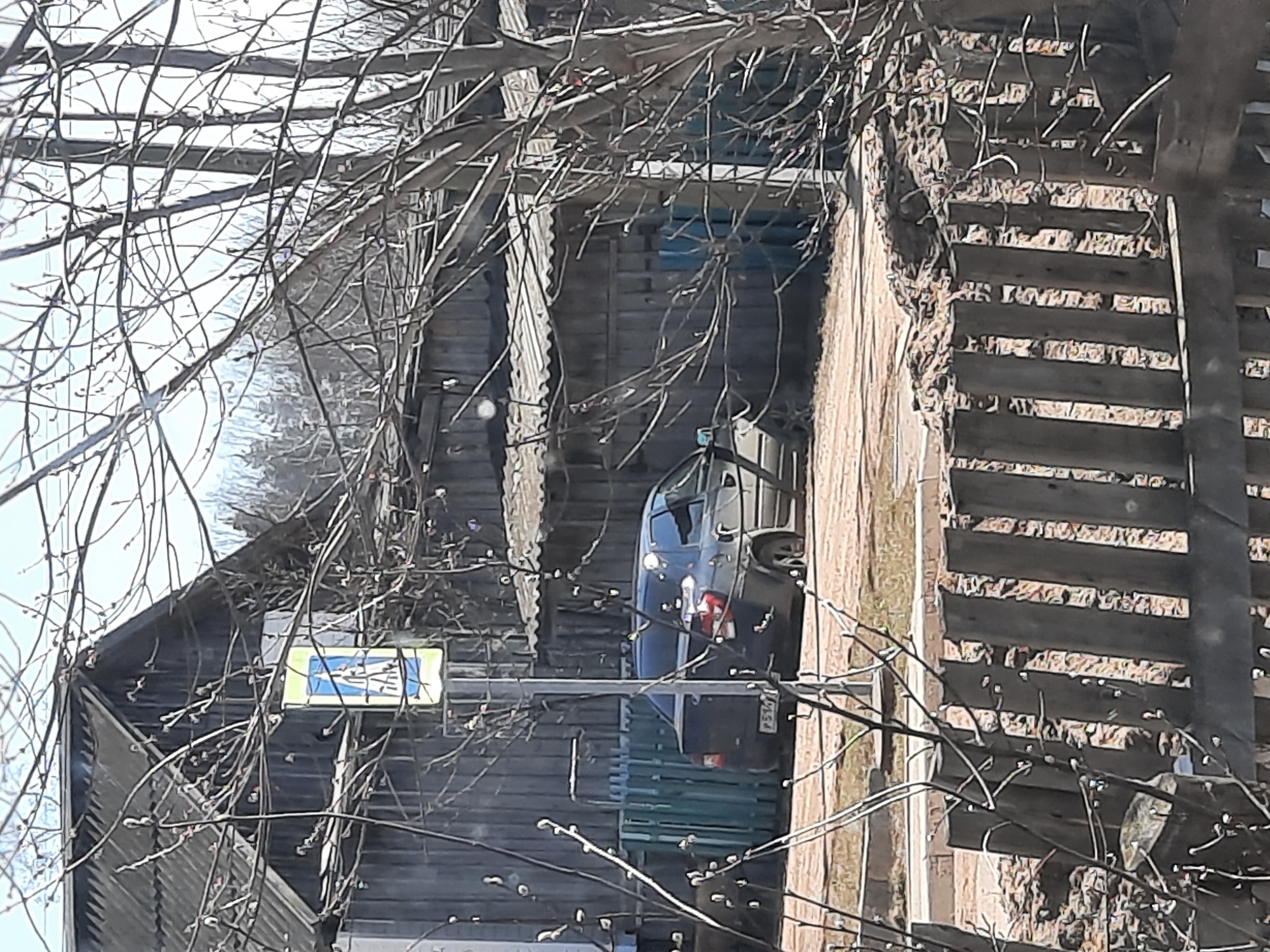 The image depicts a car parked in a narrow space between two wooden structures. The car is dark in color, and its rear license plate is partially visible. A pedestrian crossing sign is mounted on one of the wooden structures. Bare tree branches obscure portions of the view. The scene appears to be in a densely built environment, and the location is possibly in the city of Krasnogorskoye, Russia. The wooden structures appear old.