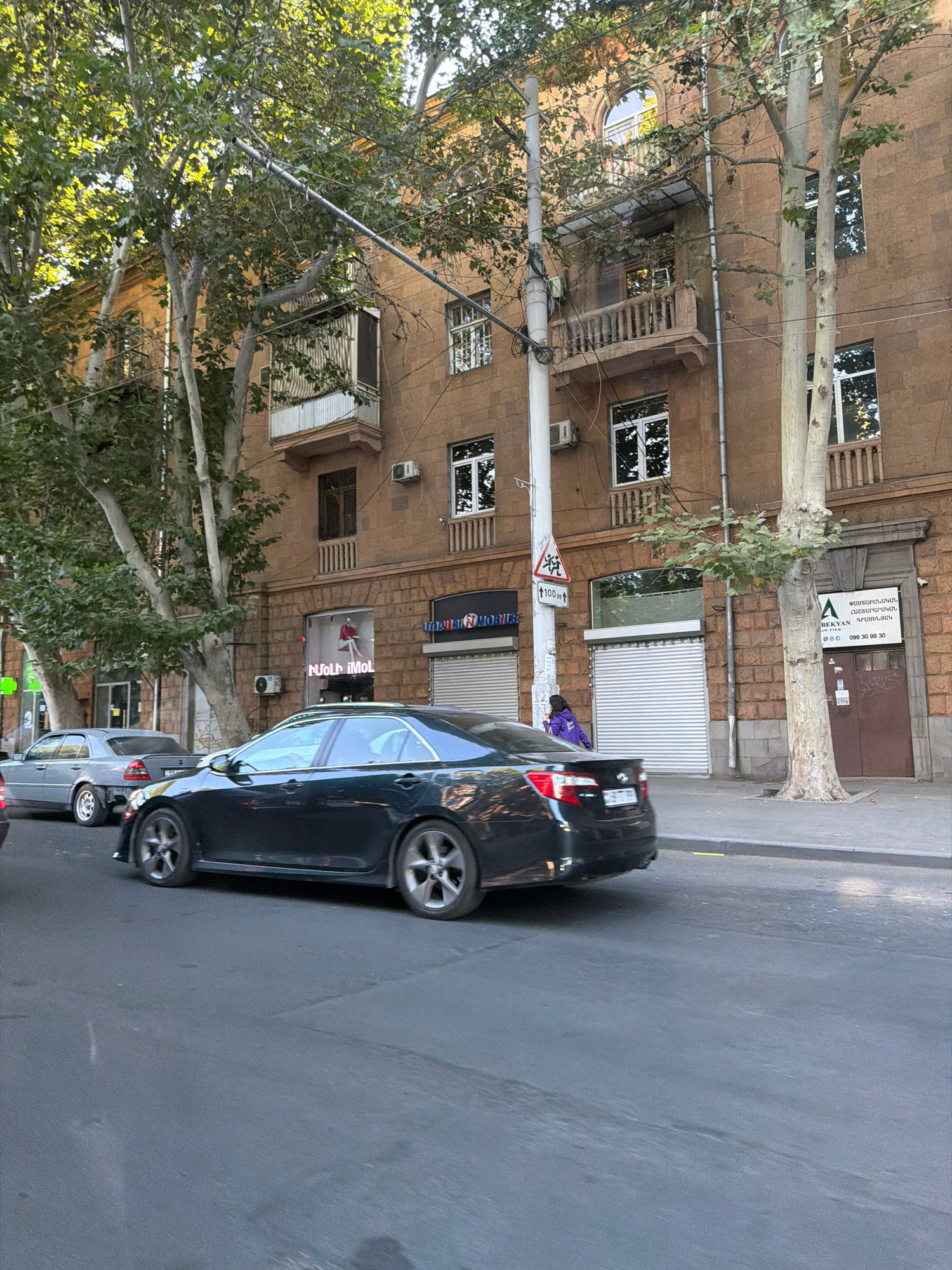 The image captures a street scene in Yerevan, Armenia, on a clear day. A dark-colored sedan is in motion on the asphalt road, its wheels partially visible. To the left, a light-colored sedan is parked, and further back, a pedestrian in a purple jacket is walking along the sidewalk. The background features a multi-story building with an ornate facade constructed of reddish-brown stone. The building has several windows, some with balconies adorned with railings and awnings. Trees with lush green foliage are interspersed throughout the scene, with their branches extending towards the building and over the street. Utility poles and wires are present, indicating urban infrastructure. Signs are visible on the building, including one with Armenian script and a phone number, and another with "100m" and a pedestrian crossing symbol. A store with a lit sign displaying "Nju i Mol" is also visible on the ground floor of the building.