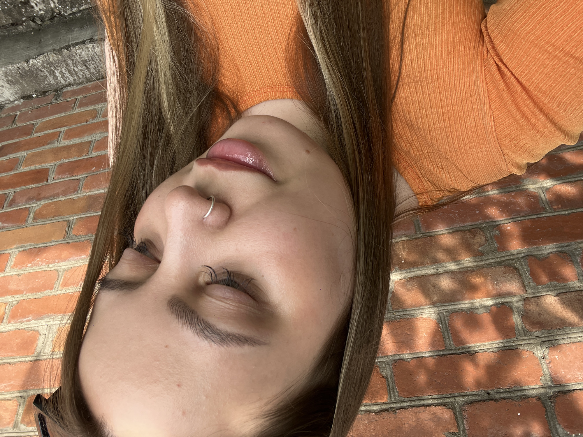 A young woman with light brown hair, wearing an orange ribbed shirt, is pictured from the chest up, looking down. She has on a silver hoop nose ring and appears to have light makeup on. Her hair is long and slightly wavy, with some lighter streaks. The background is a red brick wall, with dappled sunlight creating shadows on the bricks. The setting appears to be outdoors, with the sunlight suggesting it's daytime. The scene is devoid of any specific landmarks that would indicate the city of Novopokrovskaya, Russia.