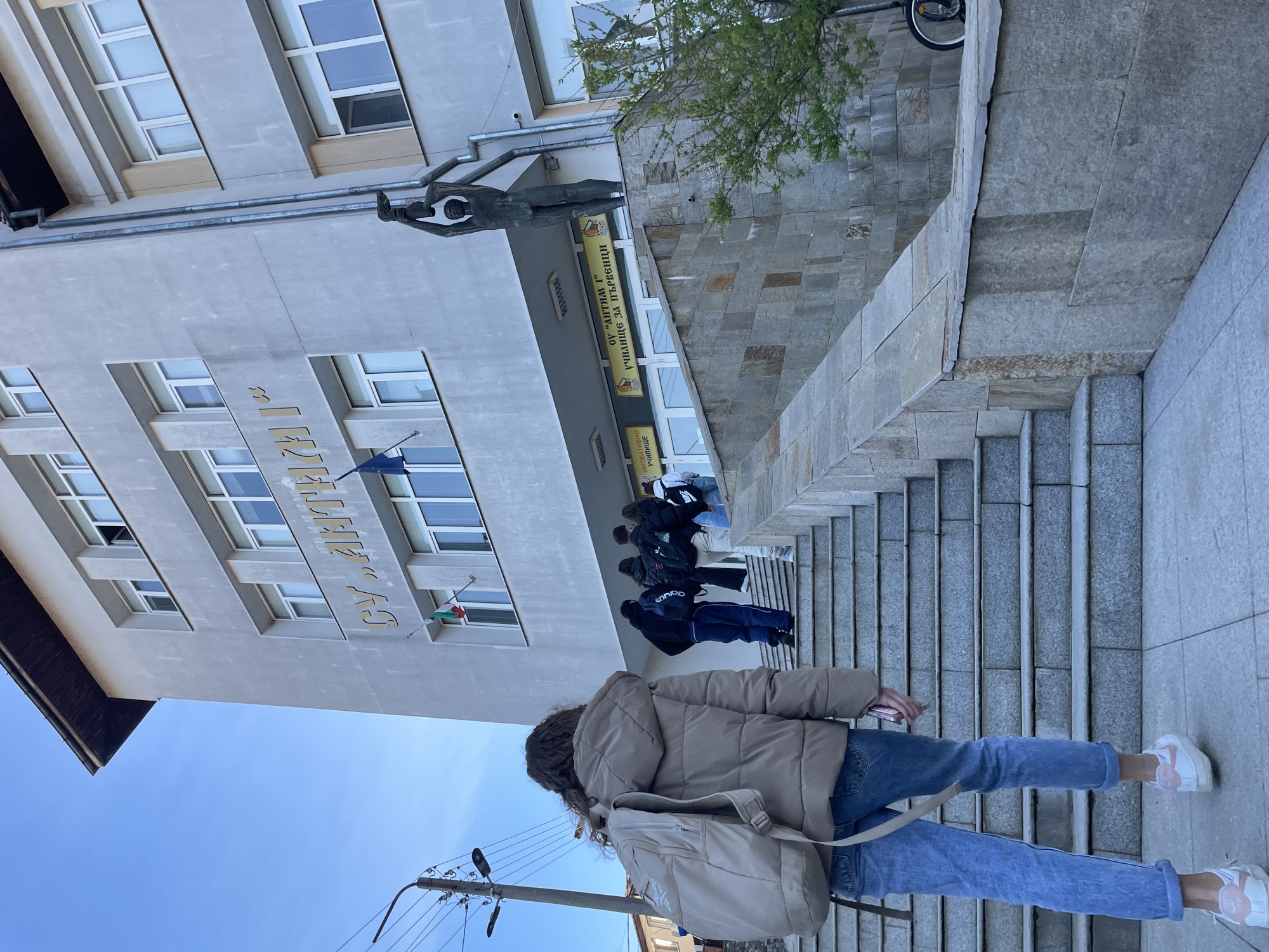 The image shows the exterior of the "SVETI KIRIL I METODIY" school in Zlatograd, Bulgaria. The school is a multi-story building with a light grey facade and numerous windows. A statue of a man stands above the entrance. Several people are walking up the steps to the entrance of the building. A person with a backpack and wearing a beige jacket and jeans walks on the steps in the foreground. A power line pole is in the lower left. The sky is visible in the upper-left.