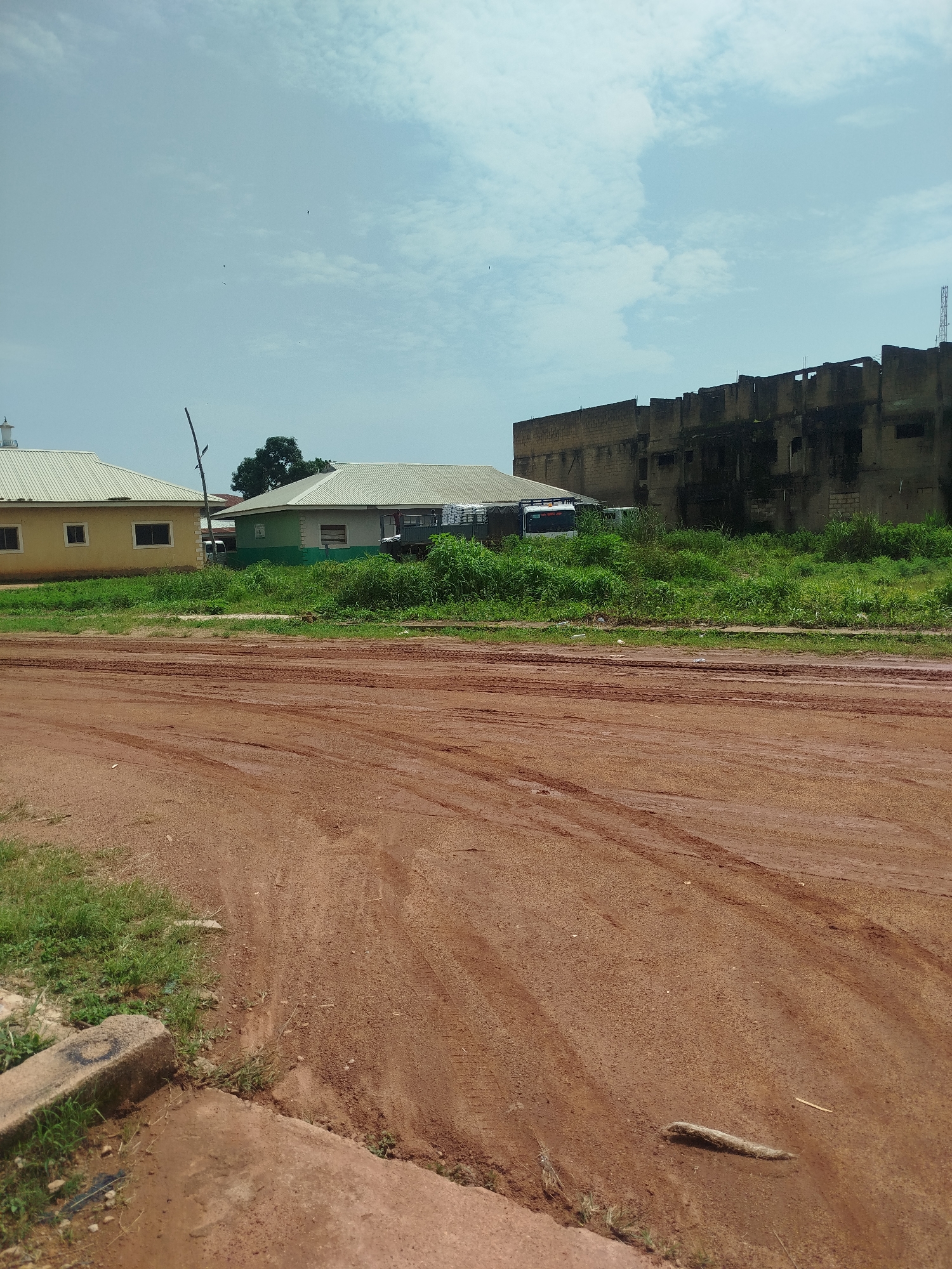 The image depicts an outdoor scene in Jos, Nigeria, dominated by a muddy, reddish-brown dirt road in the foreground, showing tire tracks. To the left, a low-slung yellow building with a corrugated metal roof and small windows is visible. Adjacent to it, a green and white building with a similar roof also stands. Behind these structures, dense green vegetation covers the ground. In the background, a large, dilapidated, multi-story building made of brick stands, its windows largely vacant and its exterior weathered and stained. The sky above is a light blue with scattered white clouds, and a few birds are visible in flight. No people are present in the image.