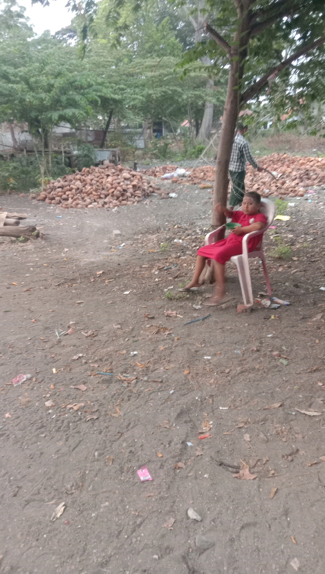 A child in a red dress sits in a white plastic chair under a tree in Karang Anyar, Indonesia. The child is holding a green bowl in their lap. A person in a checkered shirt and green pants stands behind the chair, holding a stick. The ground is covered in dirt, leaves, and scattered debris, including piles of reddish-brown objects that appear to be coconuts. In the background, trees and structures can be seen.